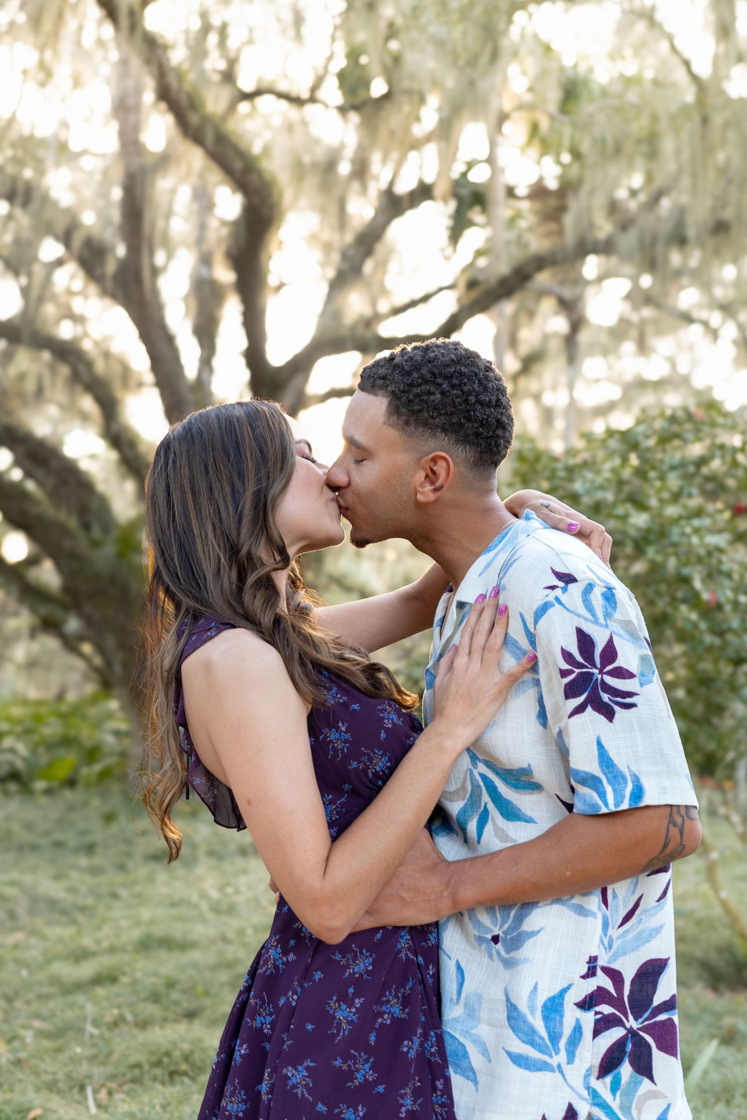 Couple kissing tenderly outdoors beneath a sprawling moss-draped oak tree in a sunlit garden.