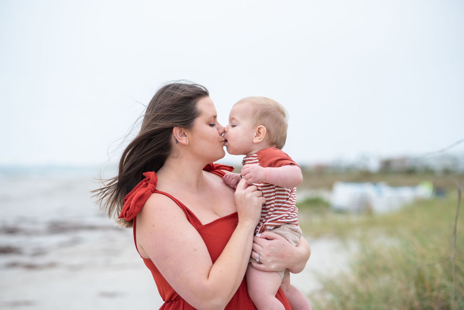 Mother in red dress kissing her baby on the beach, with ocean waves and sandy shore in the background.