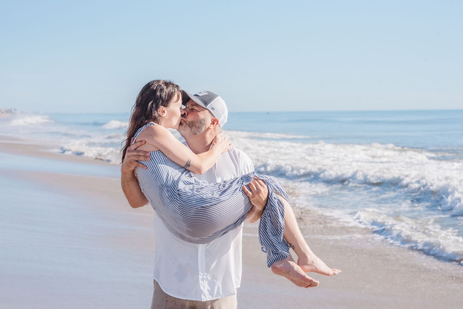 Couple sharing a romantic kiss on a sunny beach, man carrying woman in his arms near ocean waves.