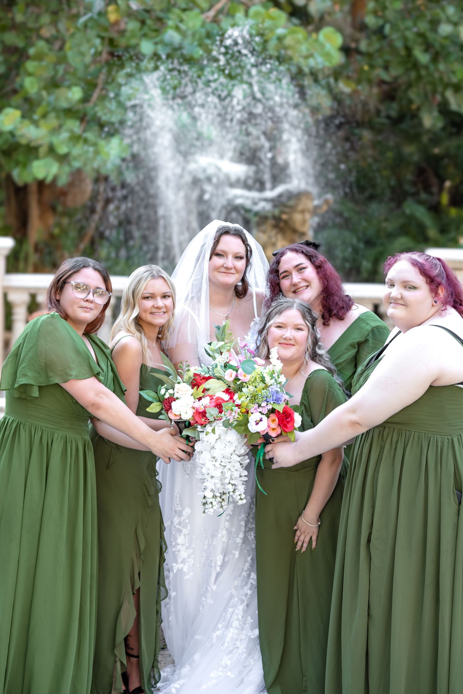 Bride in white lace gown poses with five bridesmaids in olive green dresses near a garden fountain.
