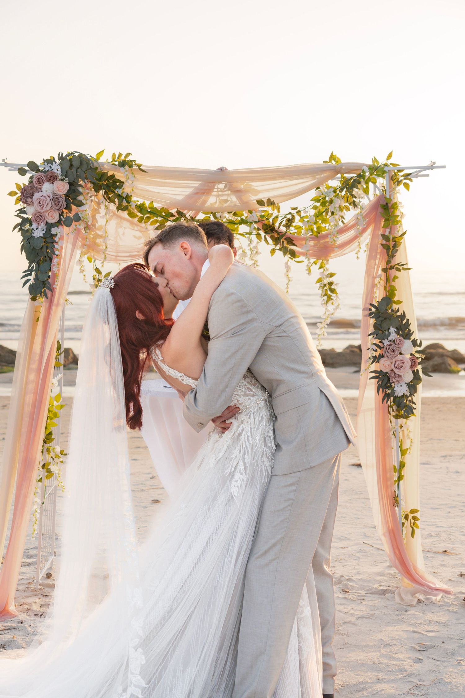 Bride and groom share first kiss under floral arch on beach at sunset wedding ceremony.