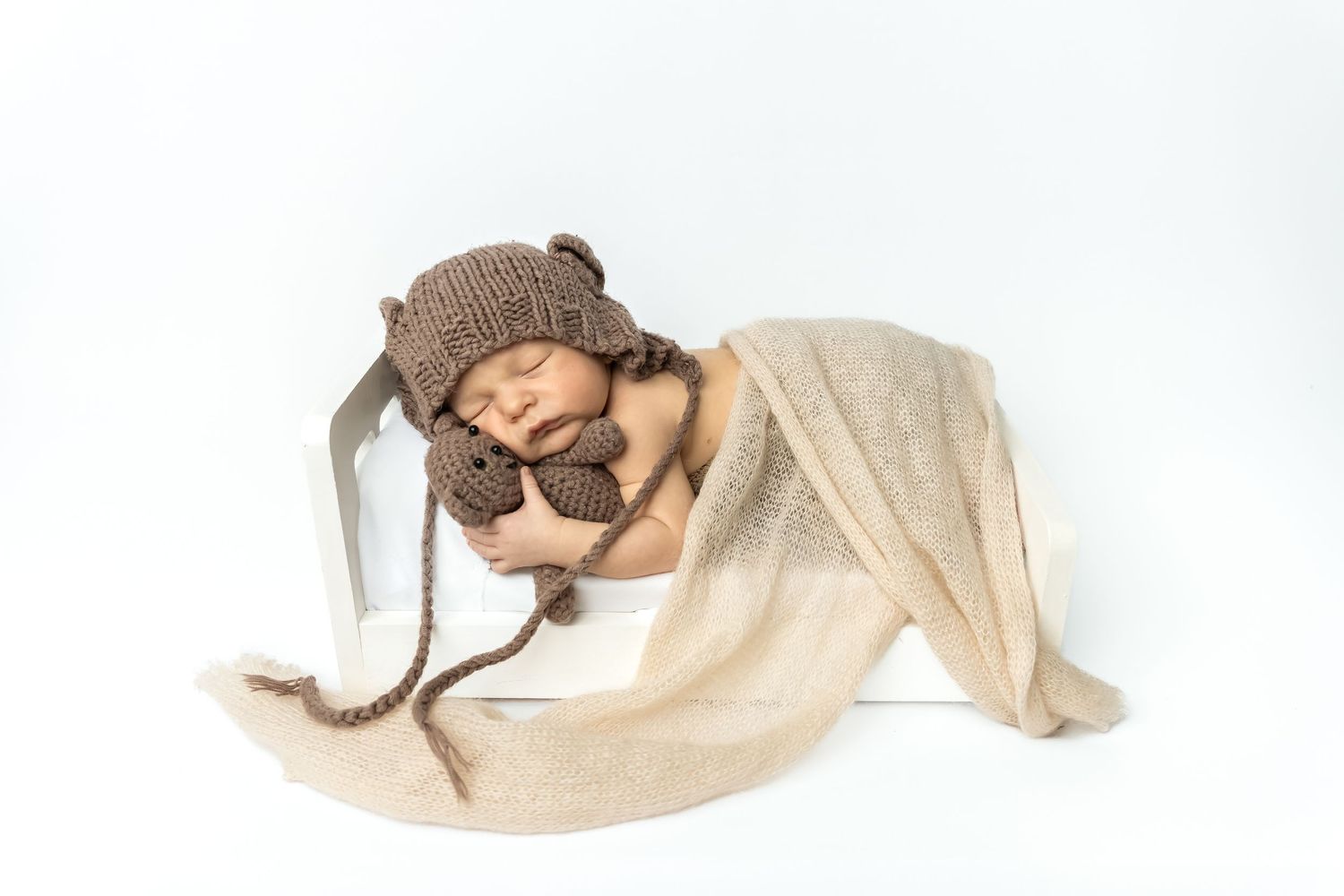 Sleeping newborn baby wearing a brown knit bear hat and booties, nestled in a white box with a beige blanket.
