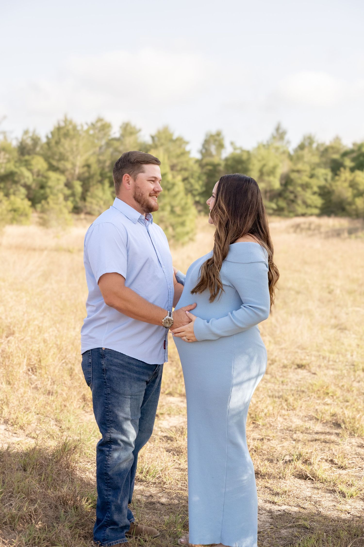 Couple in matching light blue outfits sharing a tender moment in a sunny field during a maternity photo session.