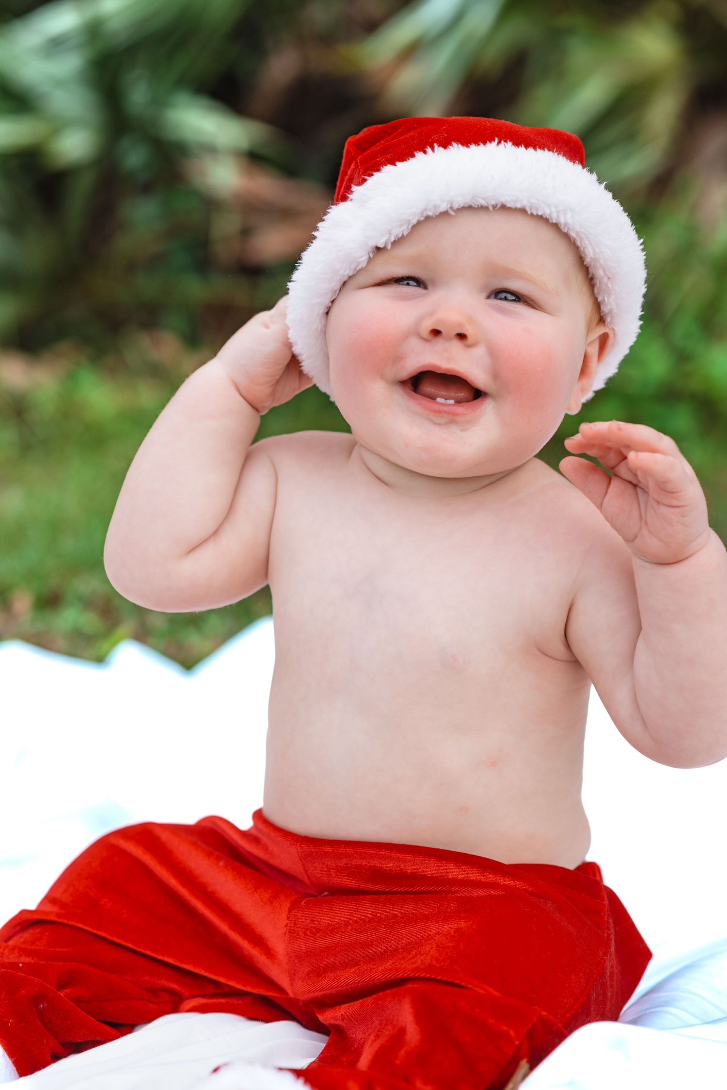 Adorable baby in Santa hat and red pants smiling outdoors for a festive Christmas photo.