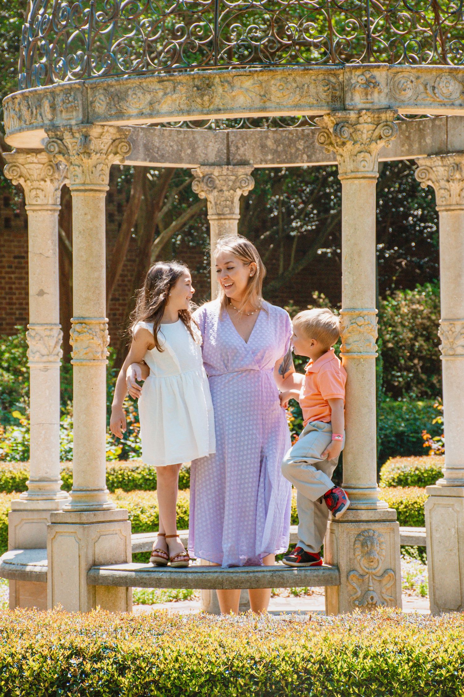 Pregnant woman in lavender dress with two children under ornate garden gazebo on sunny day.