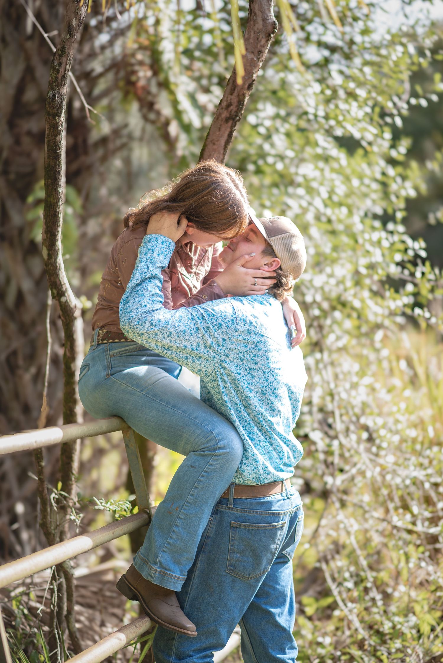 Couple embracing outdoors near rustic fence, woman in floral top and jeans, man lifting her among lush greenery.