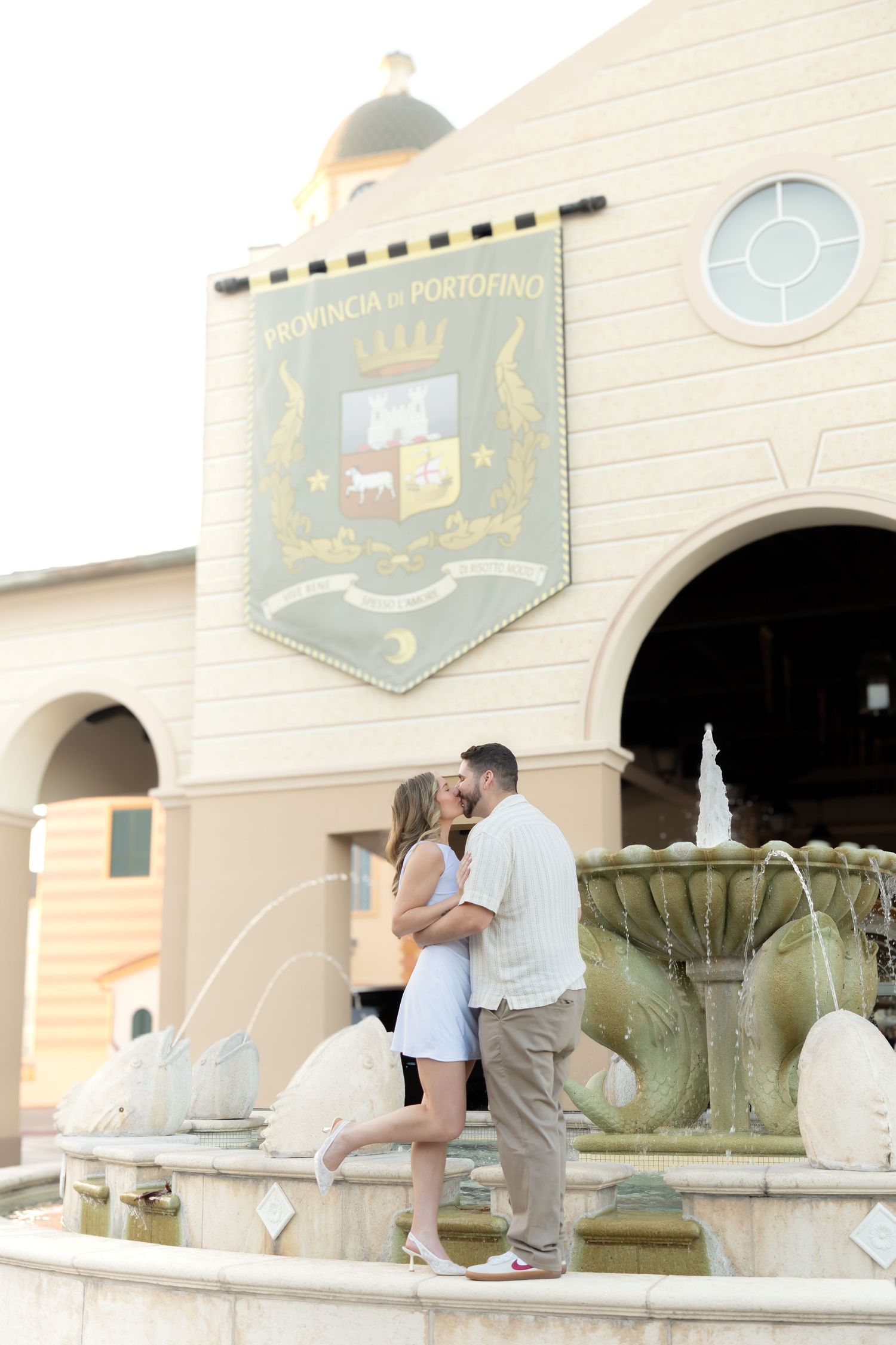 Couple kissing by a fountain at Provincia di Portofino, woman in white dress, man in khaki pants.