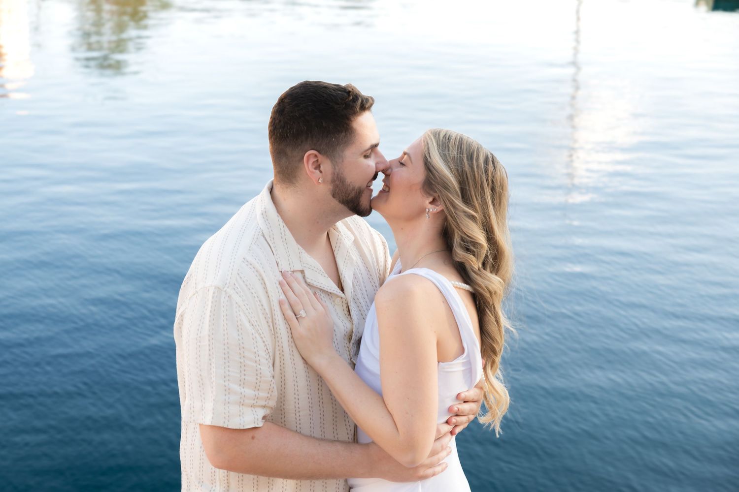 Couple sharing a romantic kiss by a serene blue waterfront, woman in white dress embracing man in cream shirt.