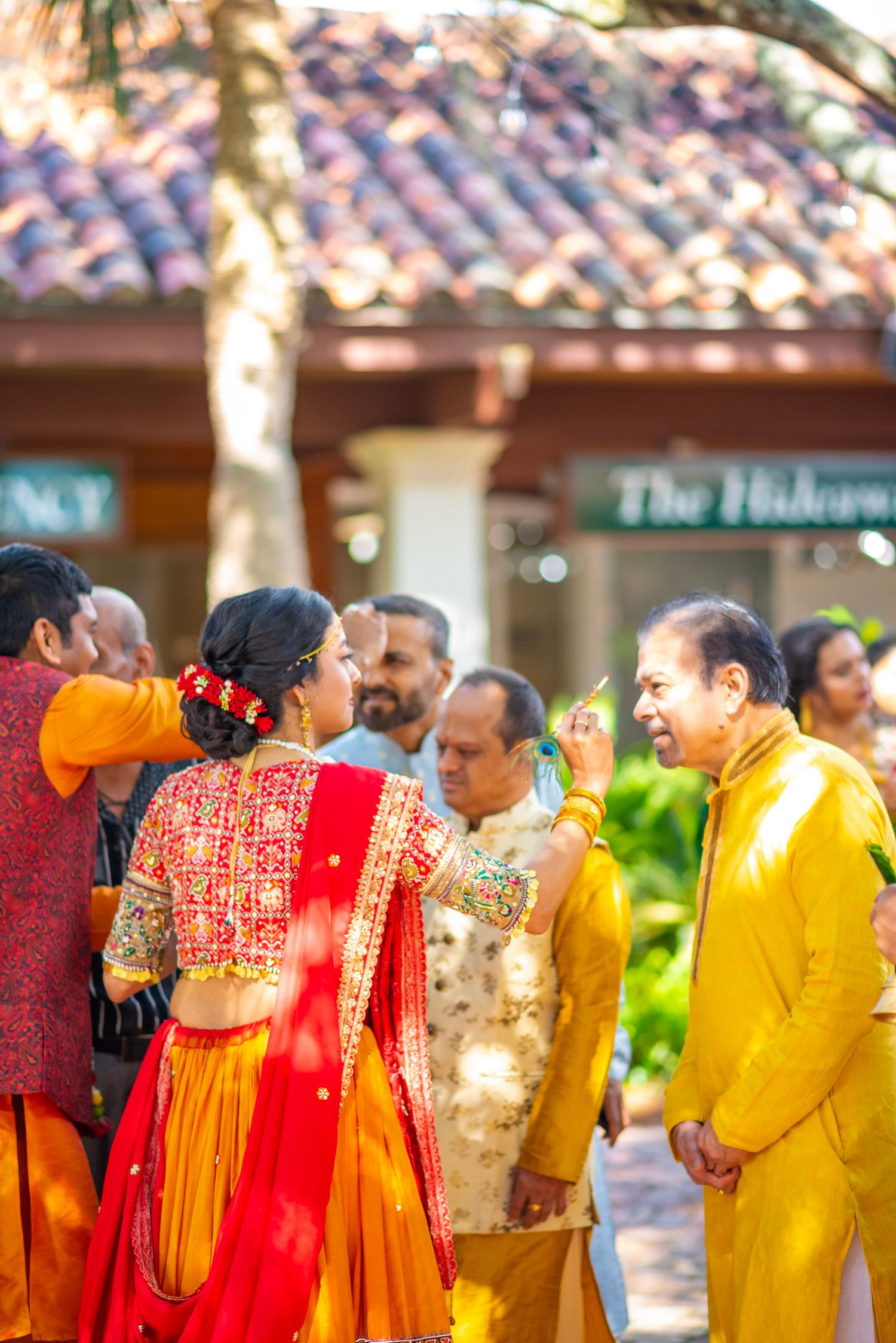 Bride in red and gold lehenga greets guests at outdoor Indian wedding celebration with colorful traditional attire.