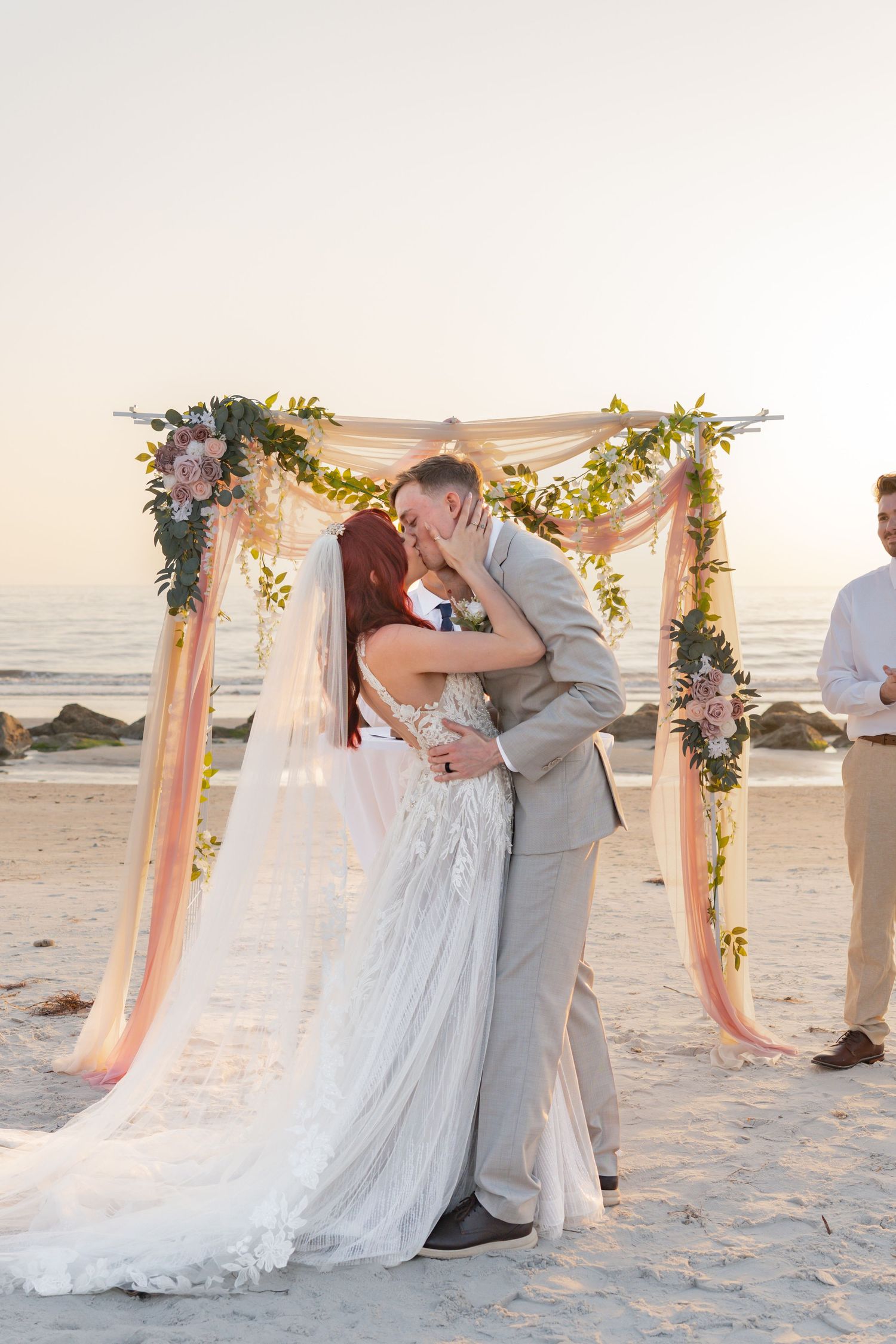 Bride and groom share first kiss under floral arch on beach at sunset during outdoor wedding ceremony.