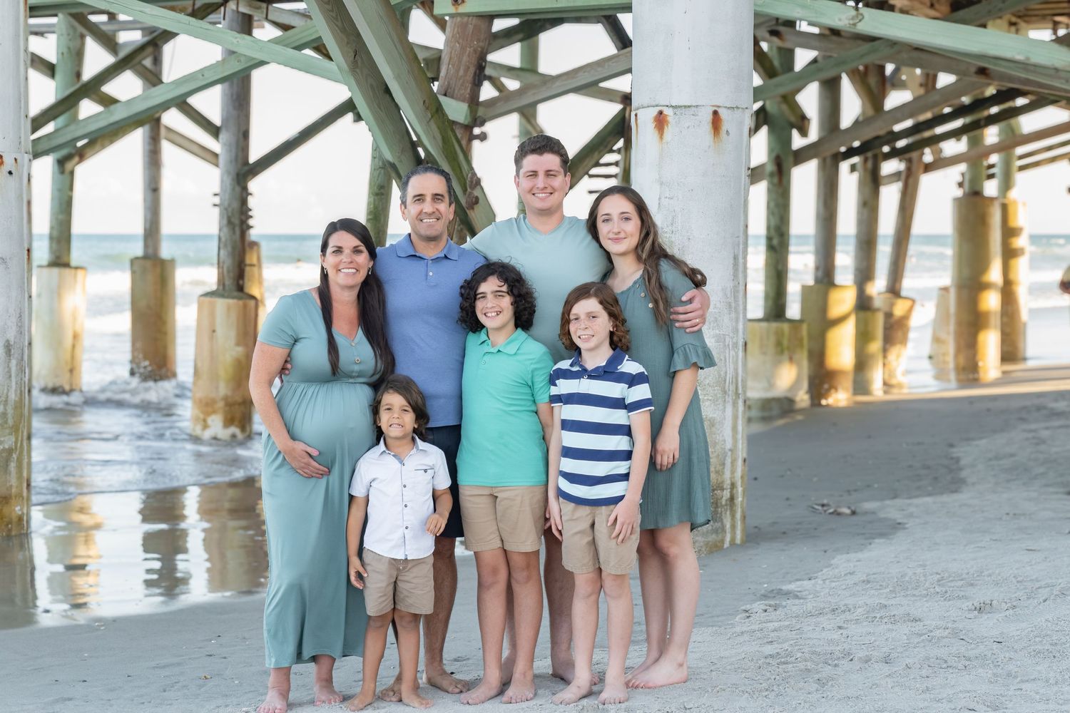 Family of seven posing together under a beach pier, with ocean waves in the background.