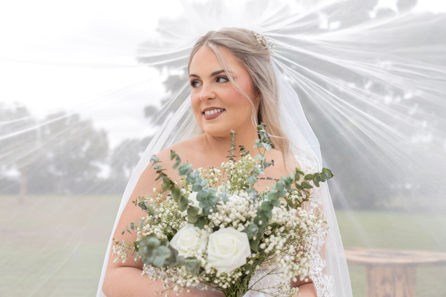 Bride holding white rose bouquet under flowing veil outdoors on wedding day.