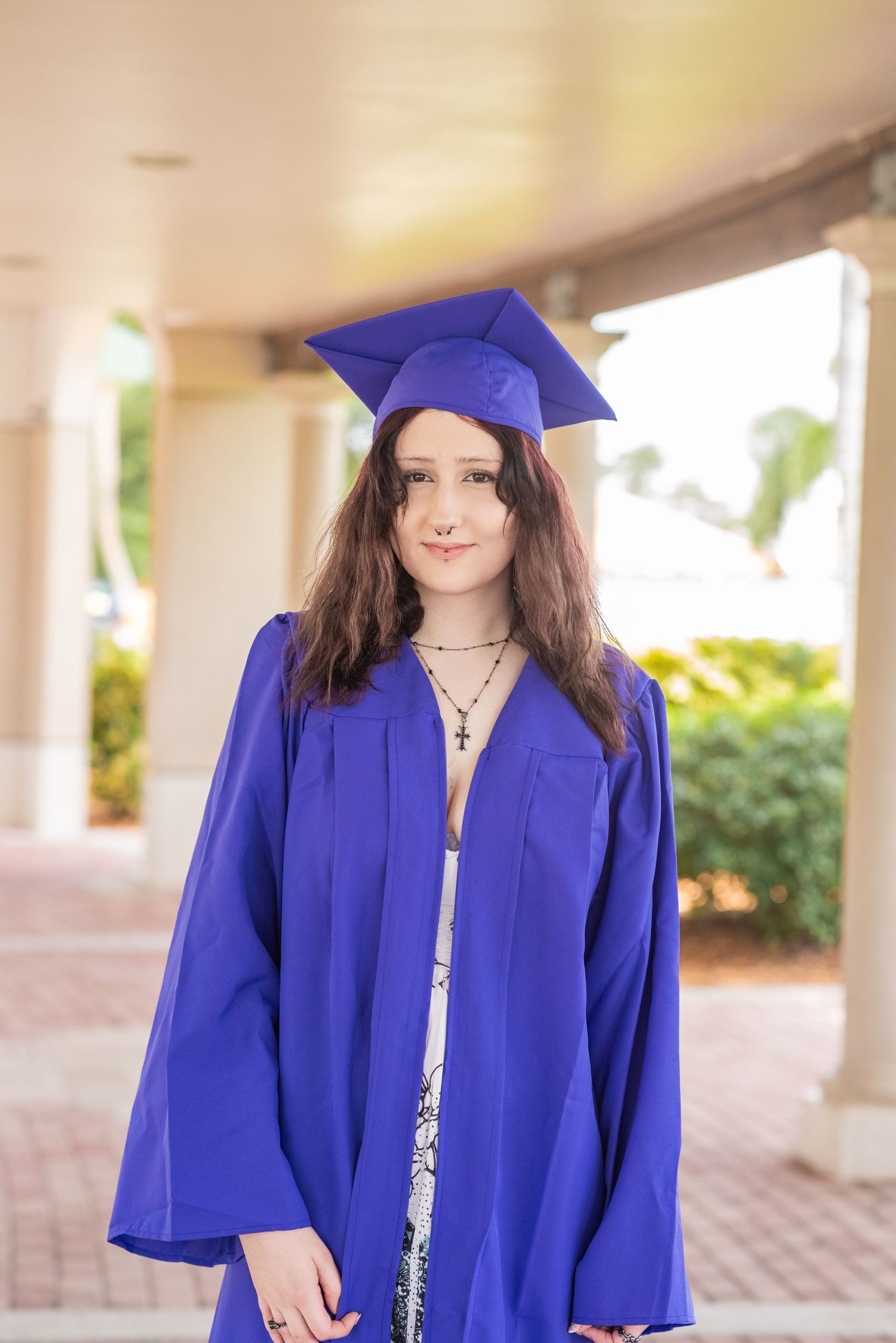 Young woman in blue graduation cap and gown smiling outdoors, celebrating academic achievement.