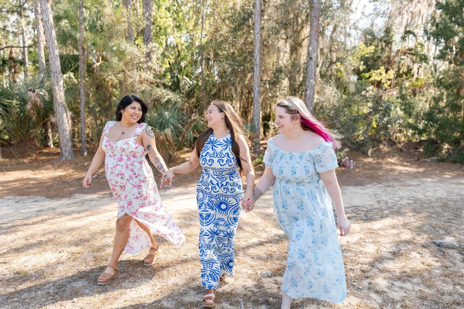 Three women in floral dresses hold hands and walk together outdoors among tall trees on a sunny day.