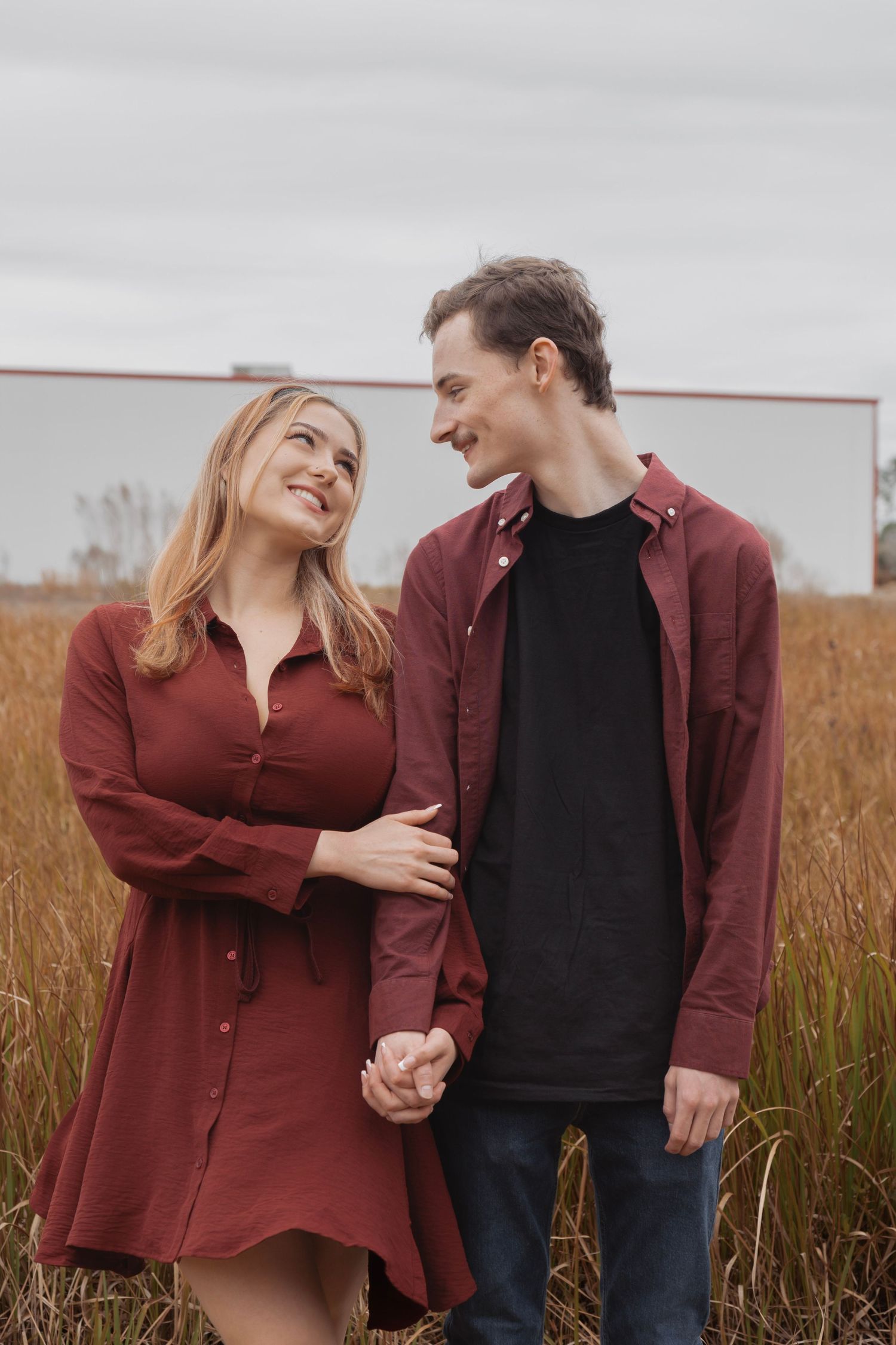 A smiling couple in matching burgundy outfits holds hands in a golden field, gazing lovingly at each other.