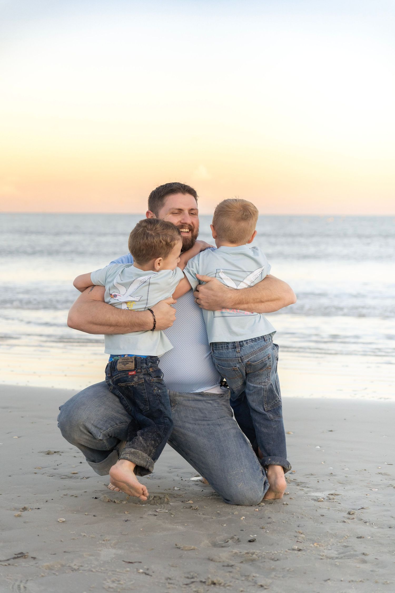 Father kneeling on sandy beach at sunset, hugging two young boys in matching outfits, ocean waves in background.