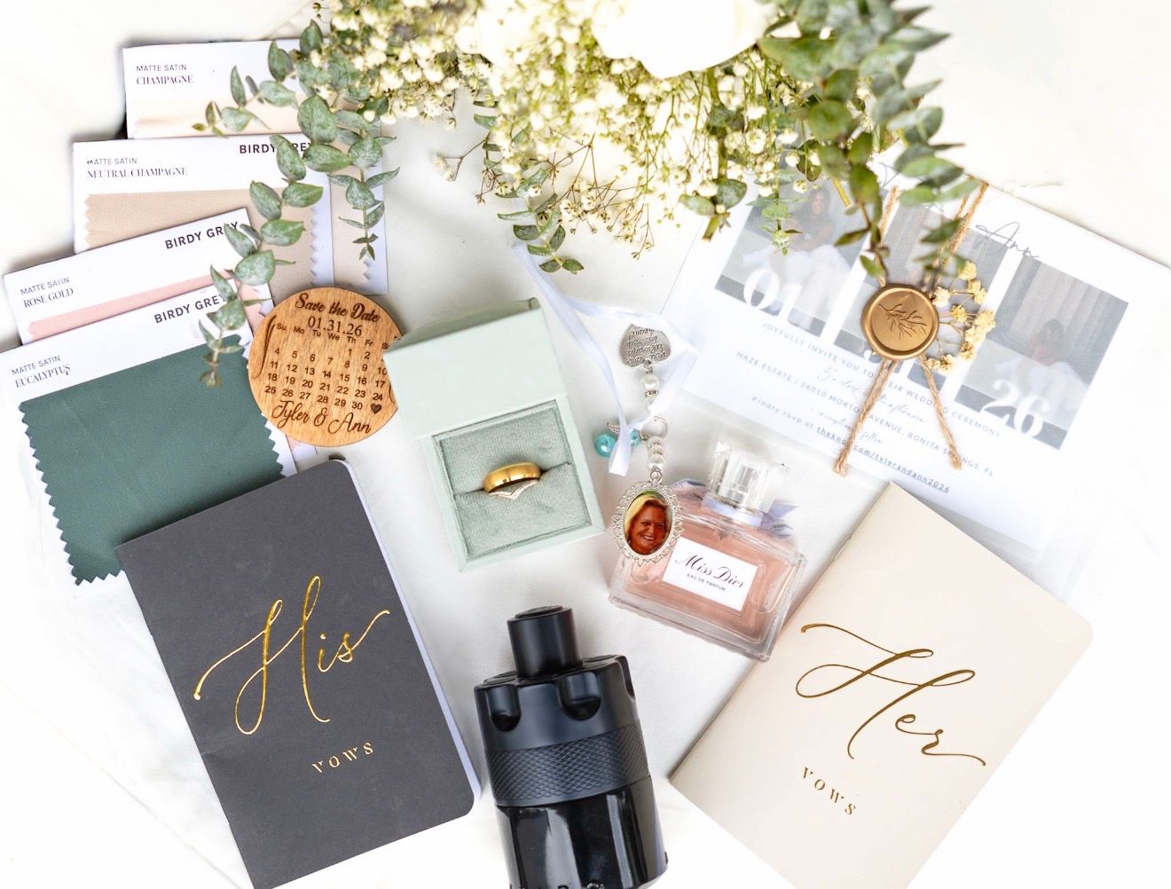 Flat lay of wedding day gifts including His and Hers vow books, ring box, perfume, and florals.