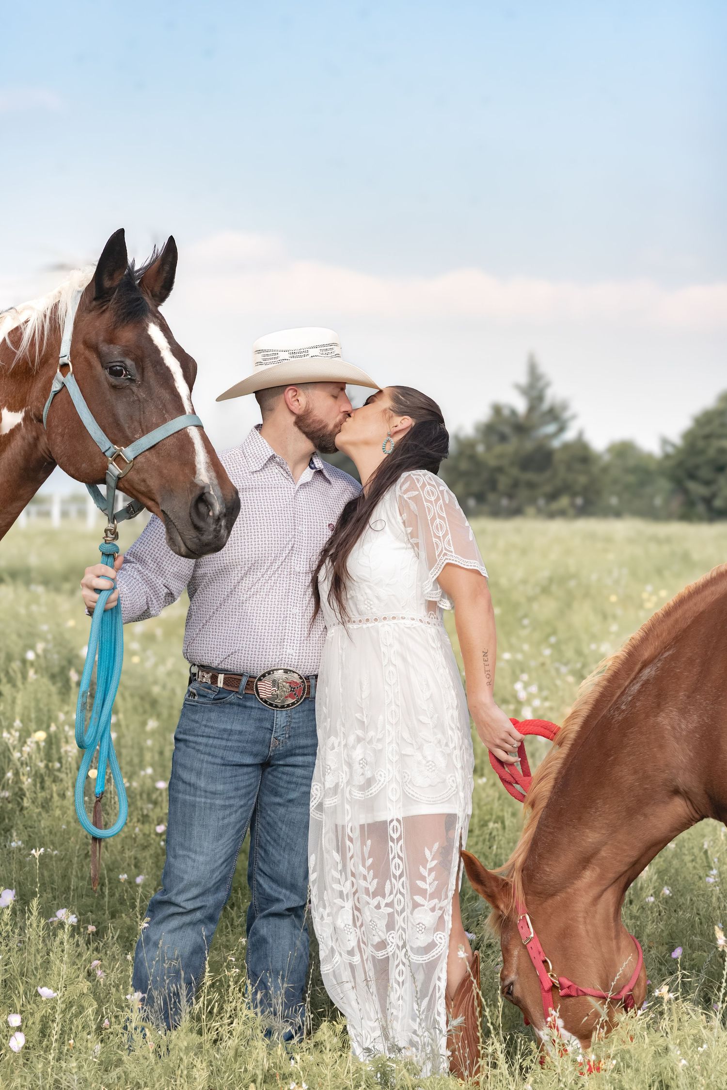 Couple shares a kiss in a wildflower field while holding horses on a beautiful sunny day.