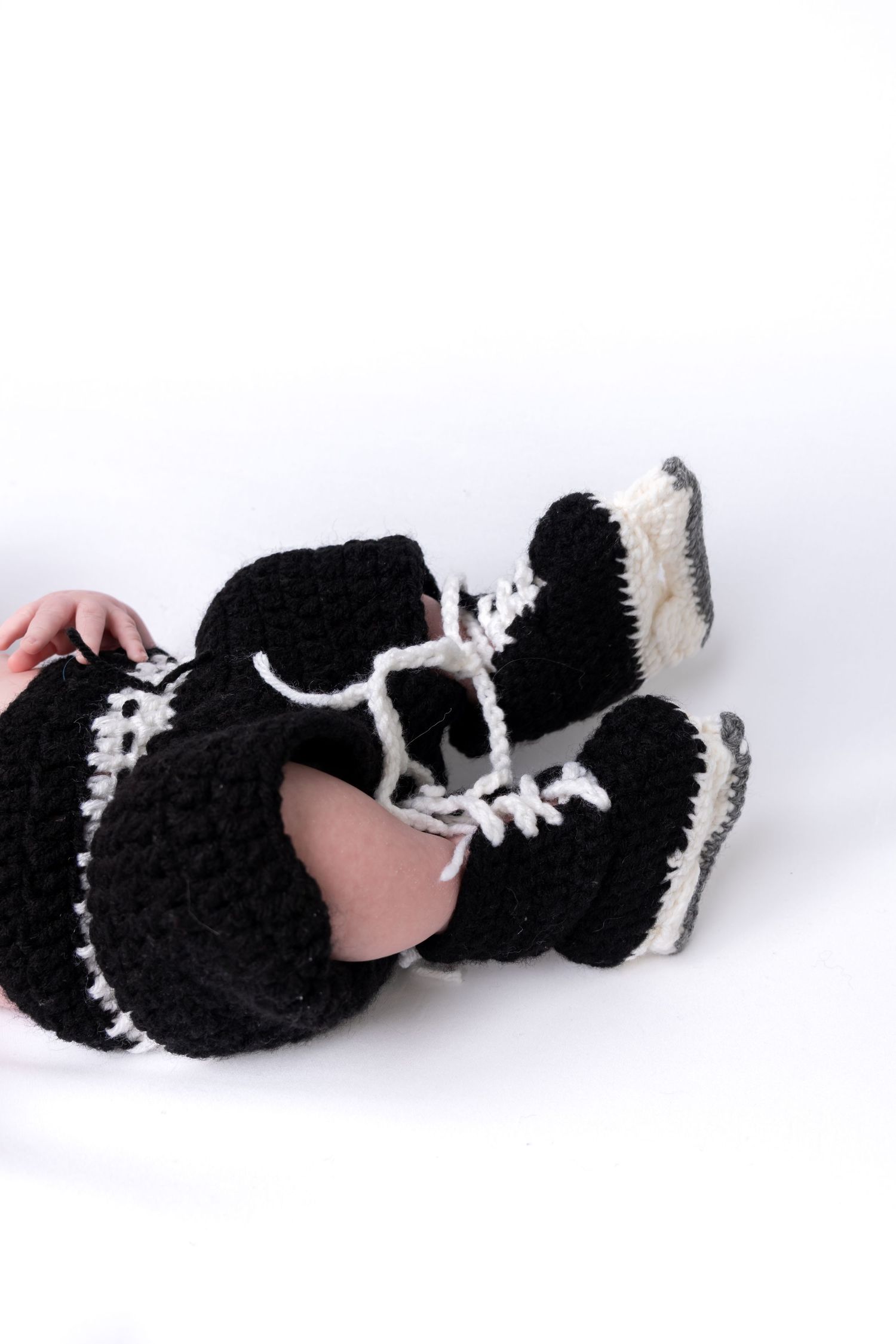 Baby wearing crocheted black and white hockey skate booties and pants costume on white background.