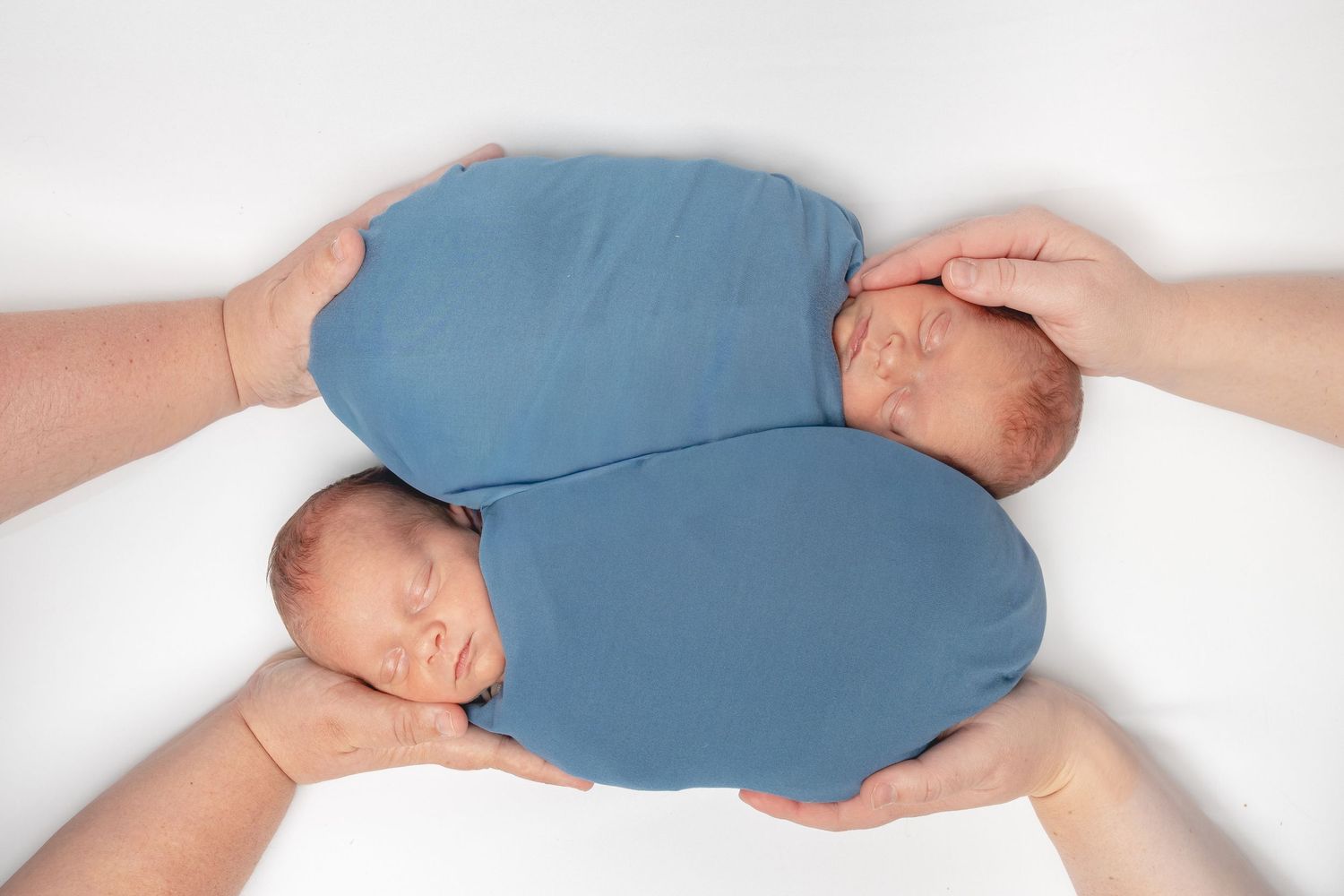Two sleeping newborn twins swaddled in blue held by adult hands on white background.