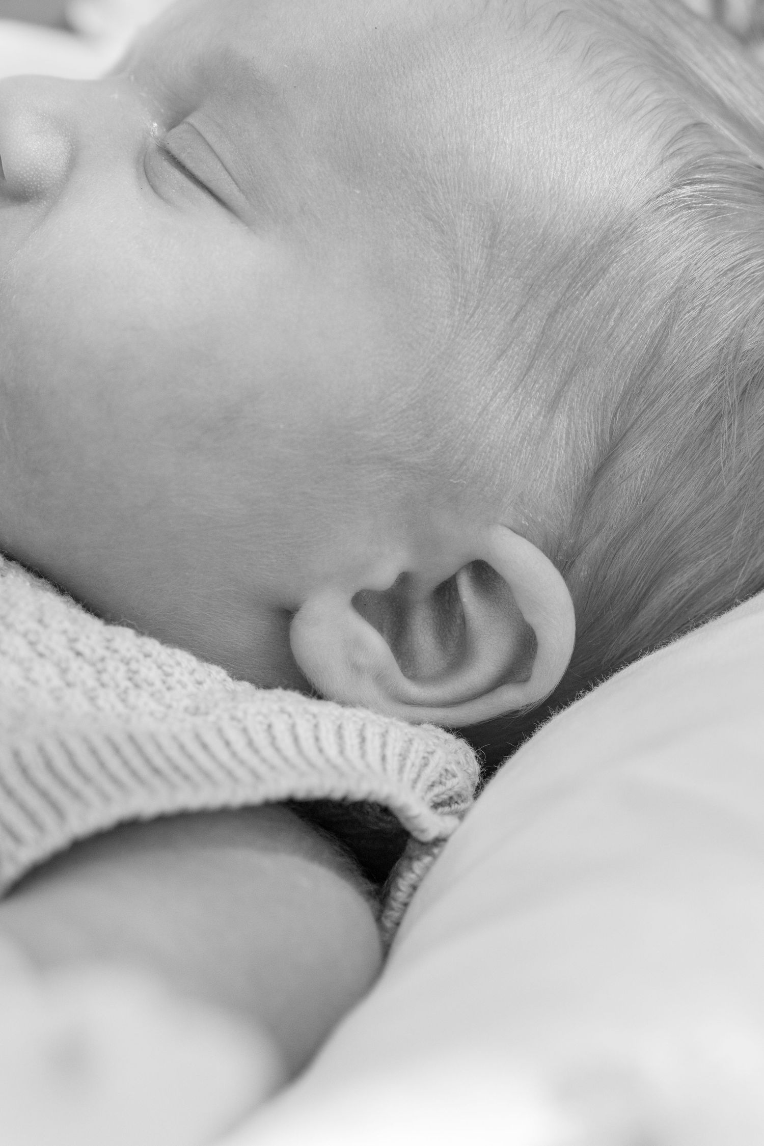 Black and white close-up photo of a sleeping newborn baby with tiny ear visible, wearing a knit sweater.