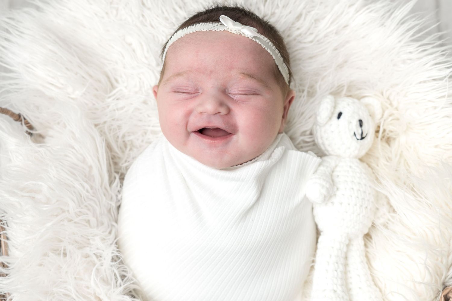 Smiling newborn baby swaddled in white, lying in fluffy basket with a knit stuffed animal toy.