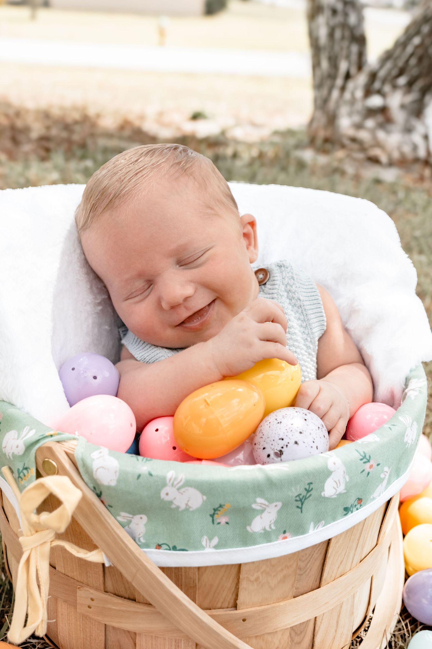Smiling newborn baby nestled in a wooden Easter basket surrounded by colorful pastel Easter eggs outdoors.