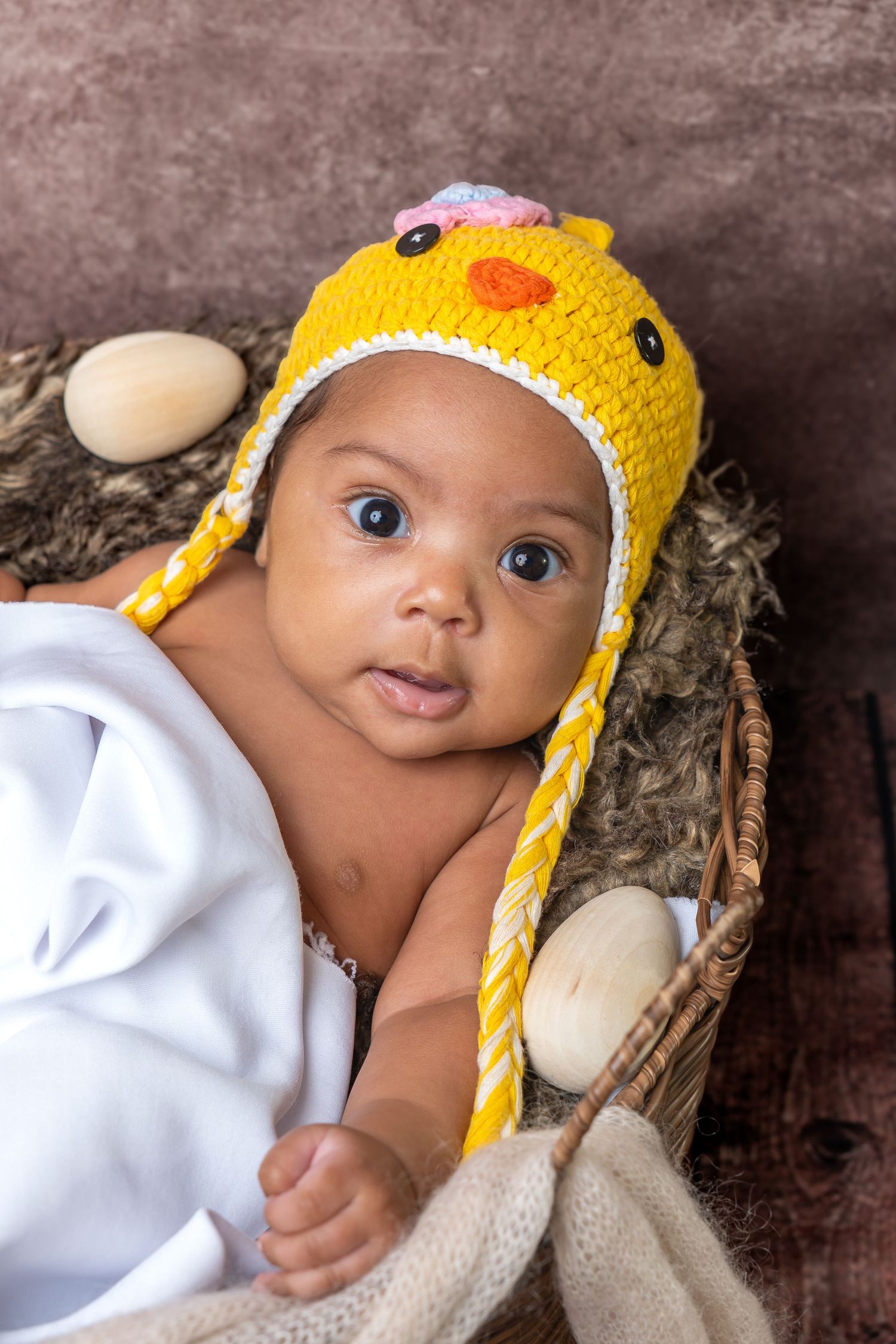 Adorable baby in yellow crocheted chick hat sitting in a basket with eggs, wrapped in white blanket.
