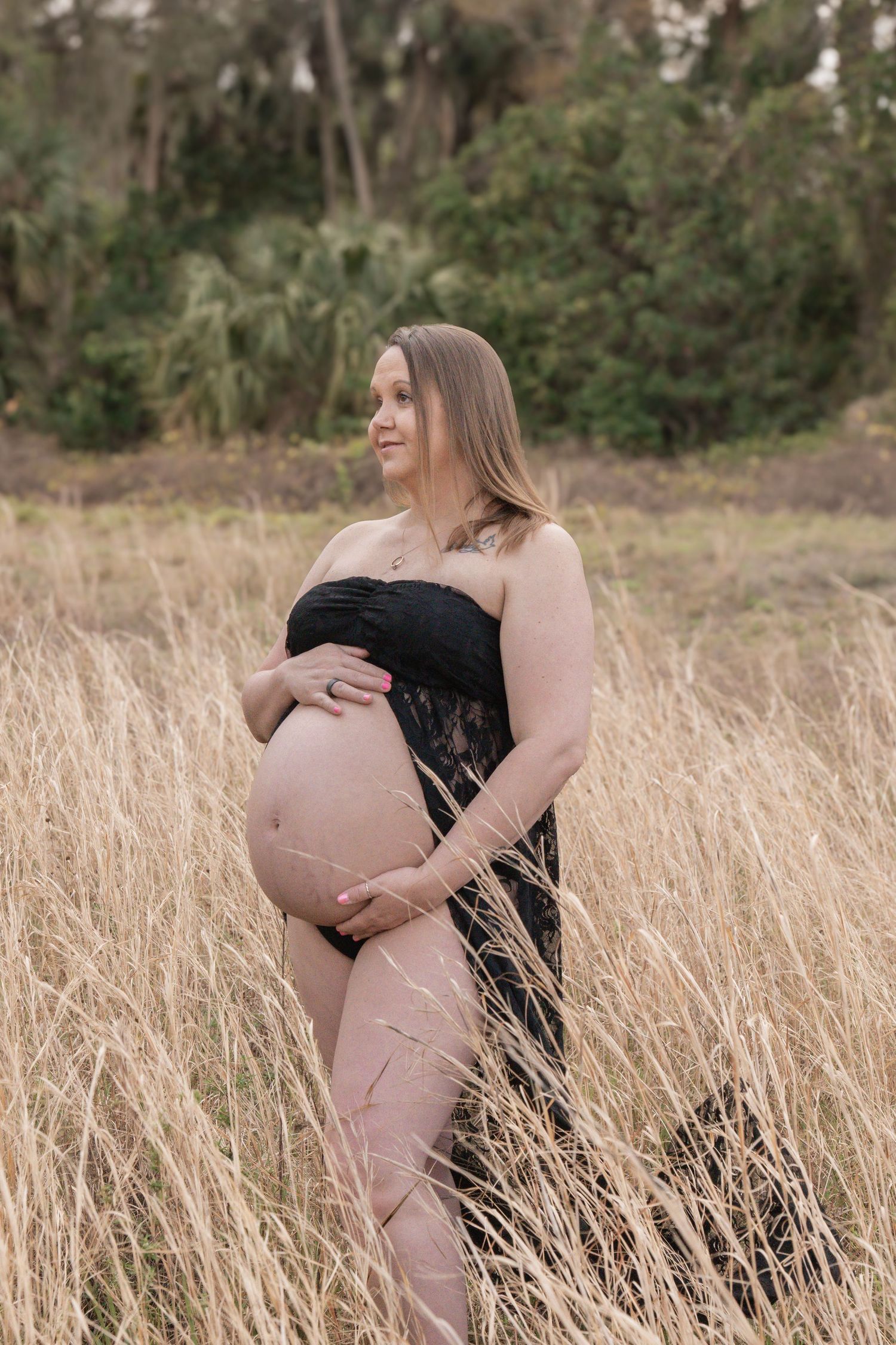 Pregnant woman in black lace wrap cradling her baby bump in a golden grass field with lush green trees behind her.