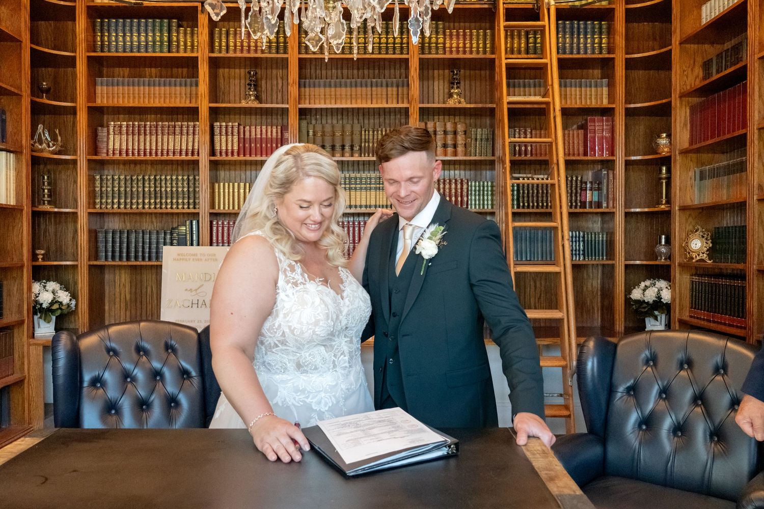 Bride and groom in a wood-paneled library review their wedding guest book together at a table.