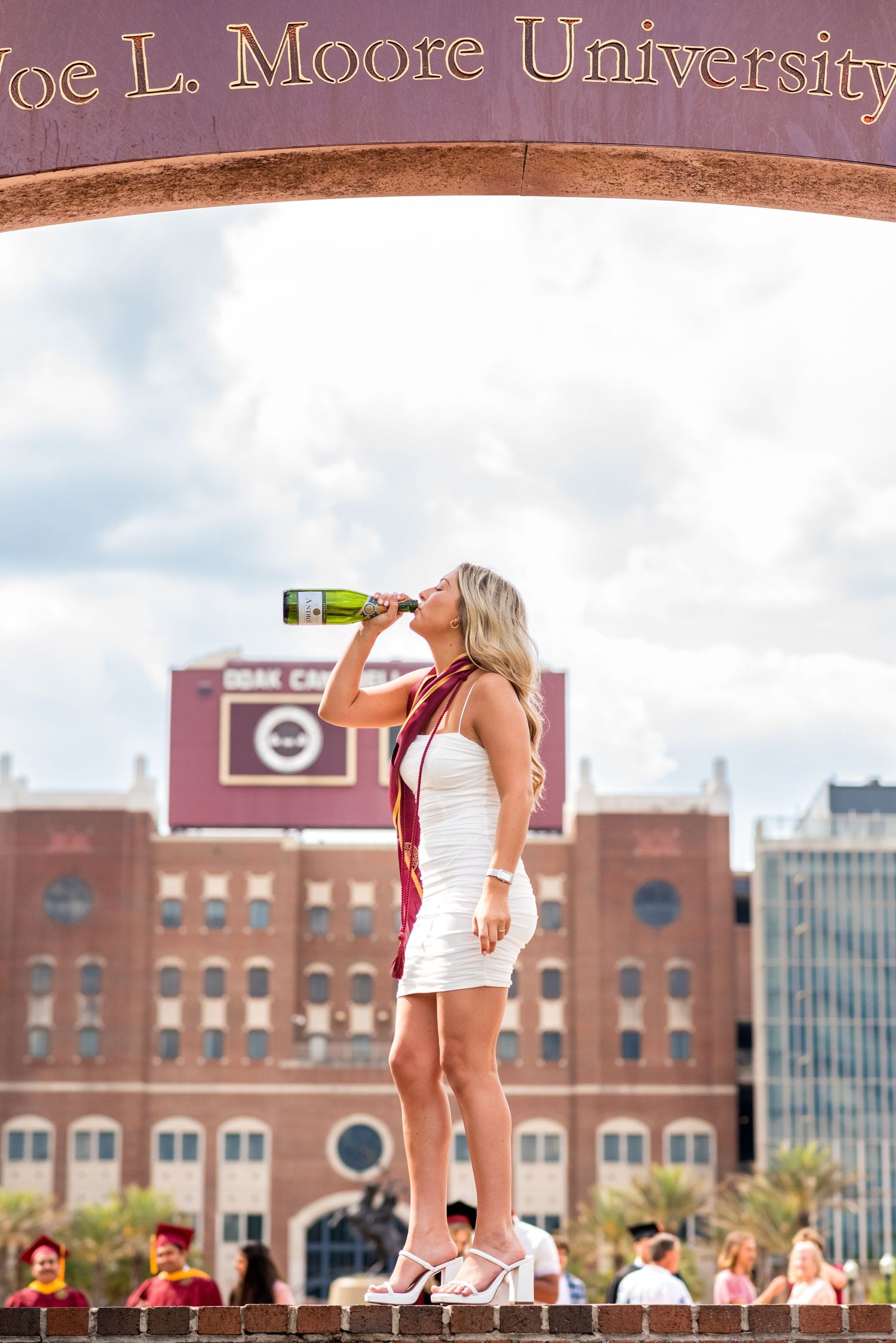 Female FSU graduate in white dress drinks champagne under Joe L. Moore University arch at Doak Campbell Stadium.