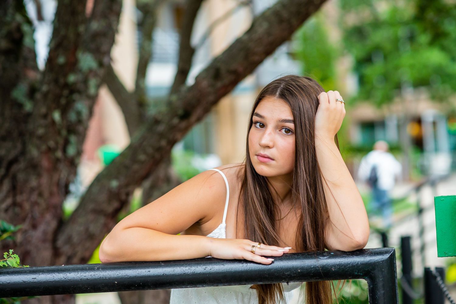 Young woman with long brown hair leaning on a railing in an urban park setting.