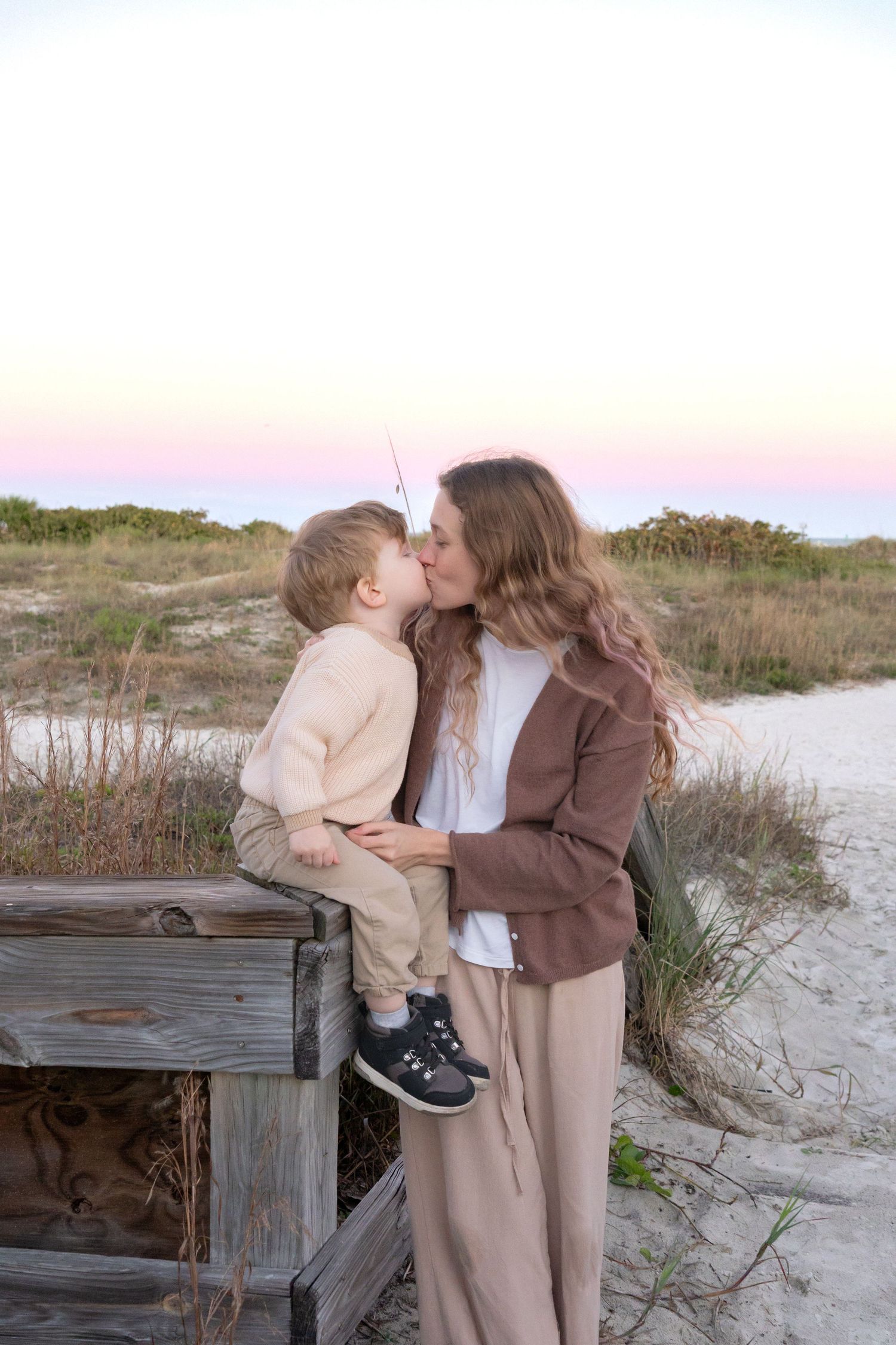 A woman kisses a young toddler boy sitting on a wooden fence at a sandy beach path during sunset.
