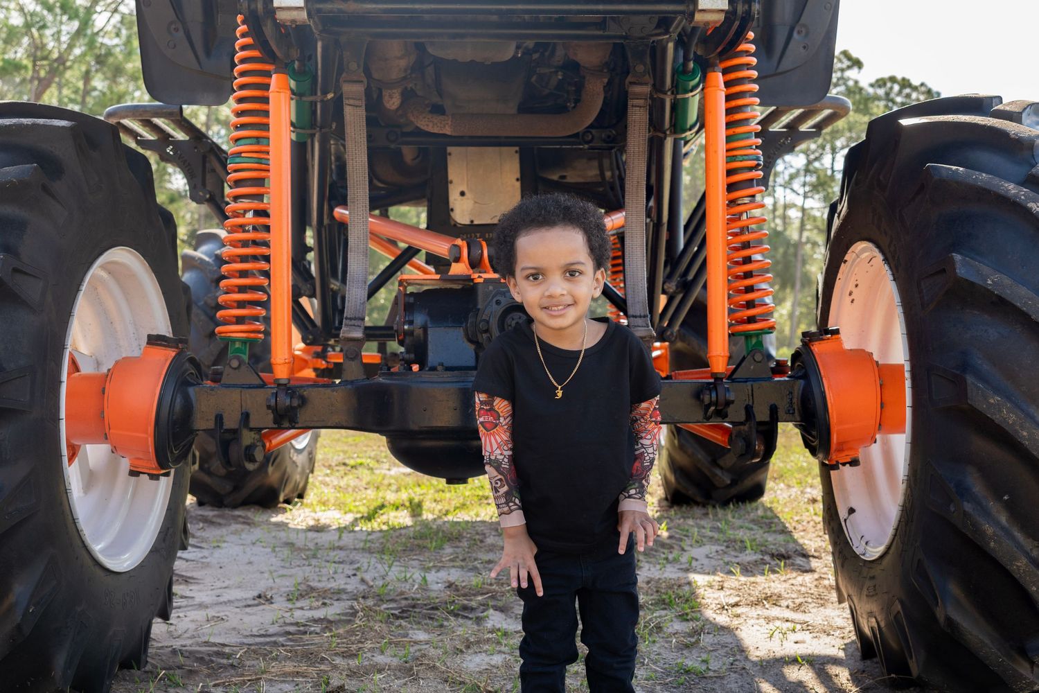 Young boy smiling and standing in front of a large orange and black monster truck with giant tires outdoors.