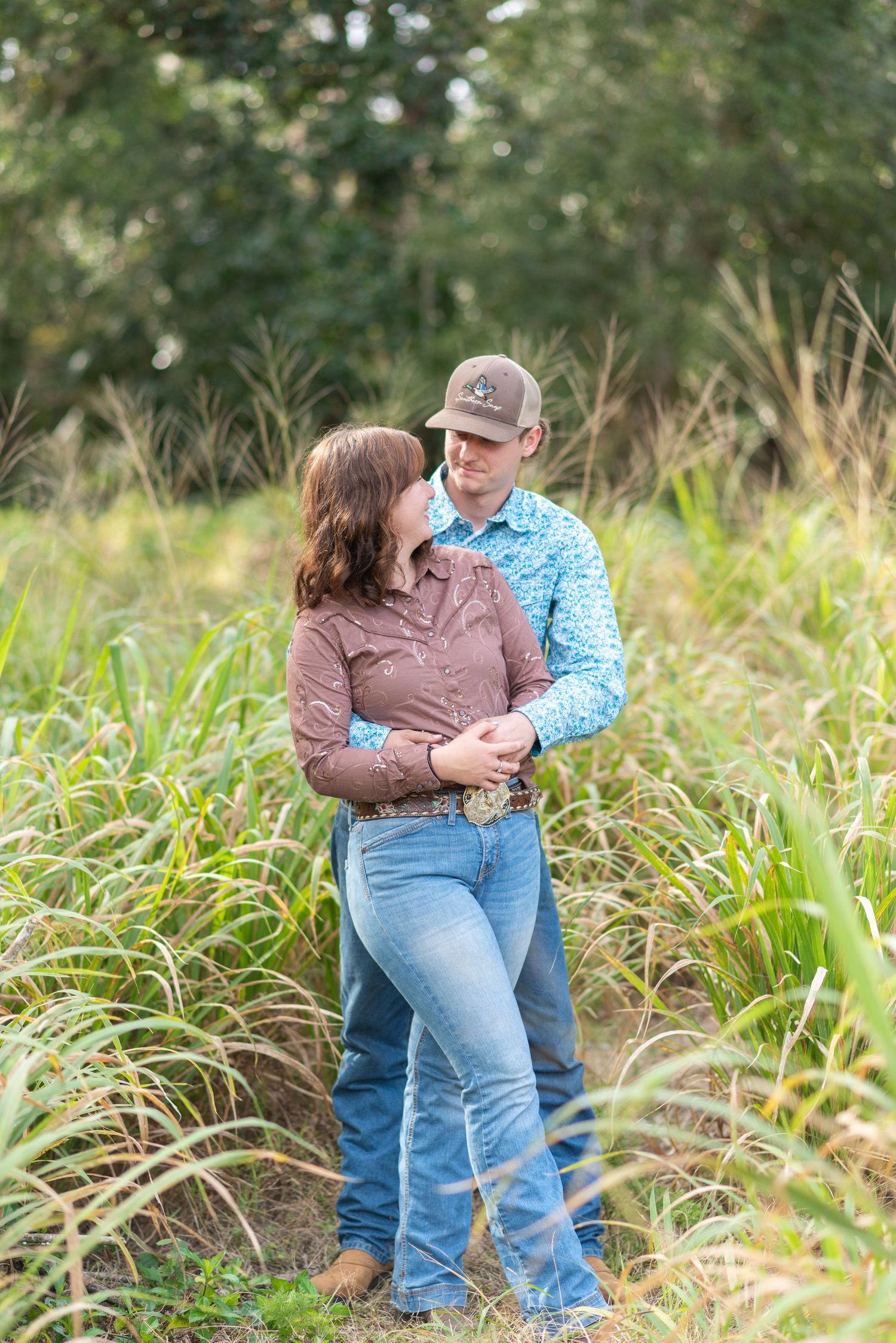 Couple embracing in a lush green field, wearing western attire including jeans and a baseball cap.