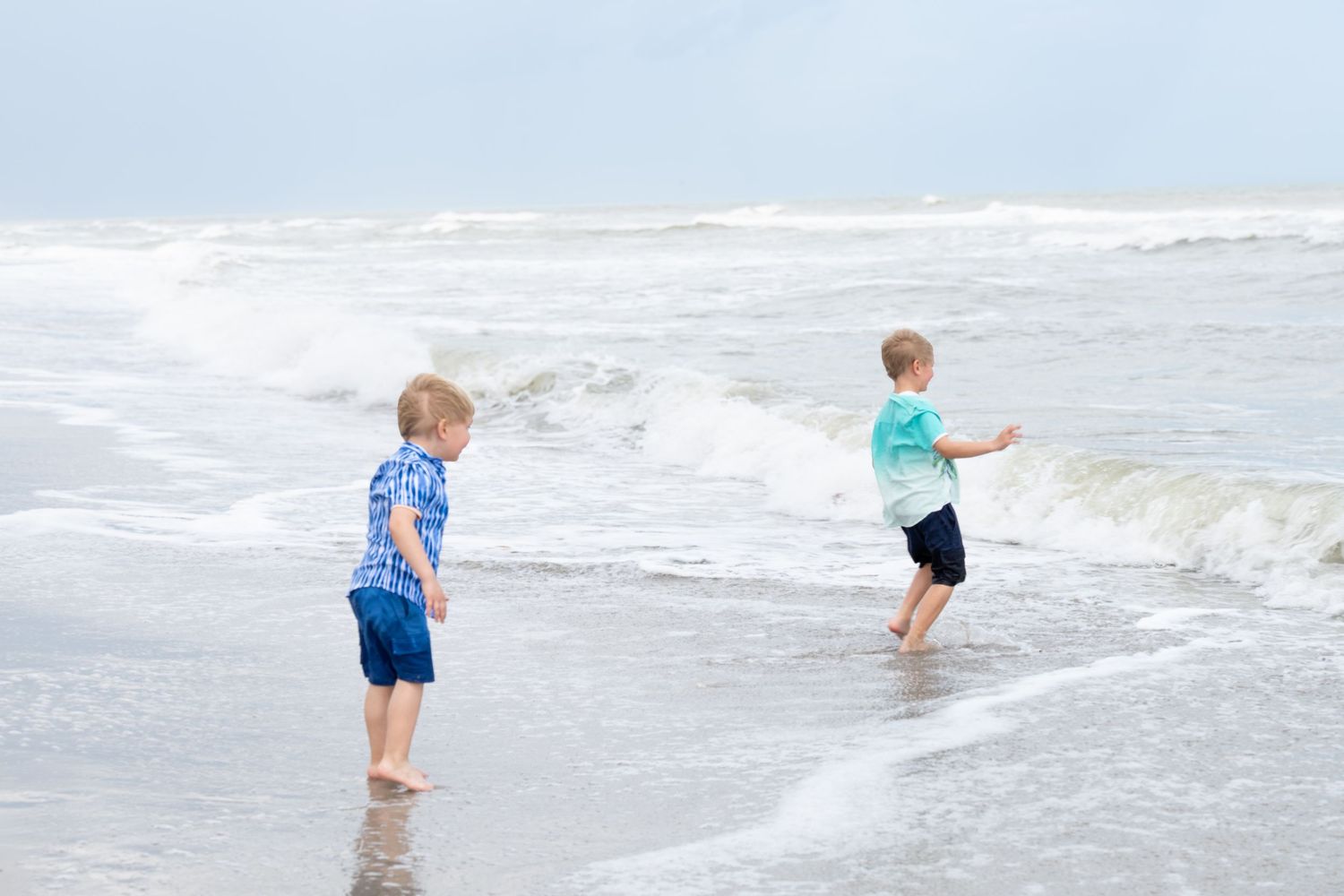 Two young blonde boys play in the ocean waves on a sandy beach on a cloudy day.