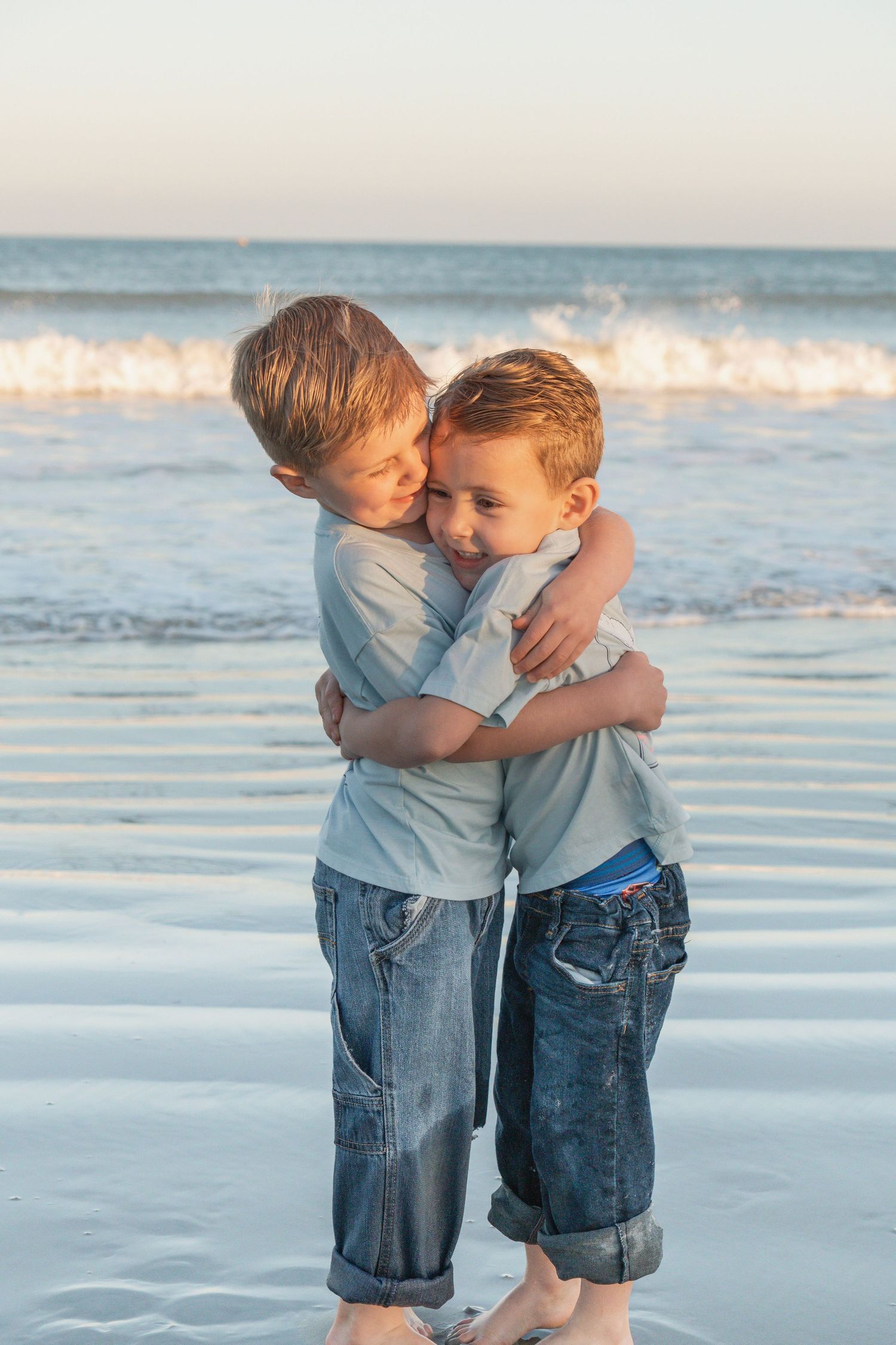 Two young boys hugging each other on a sandy beach with ocean waves in the background at sunset.