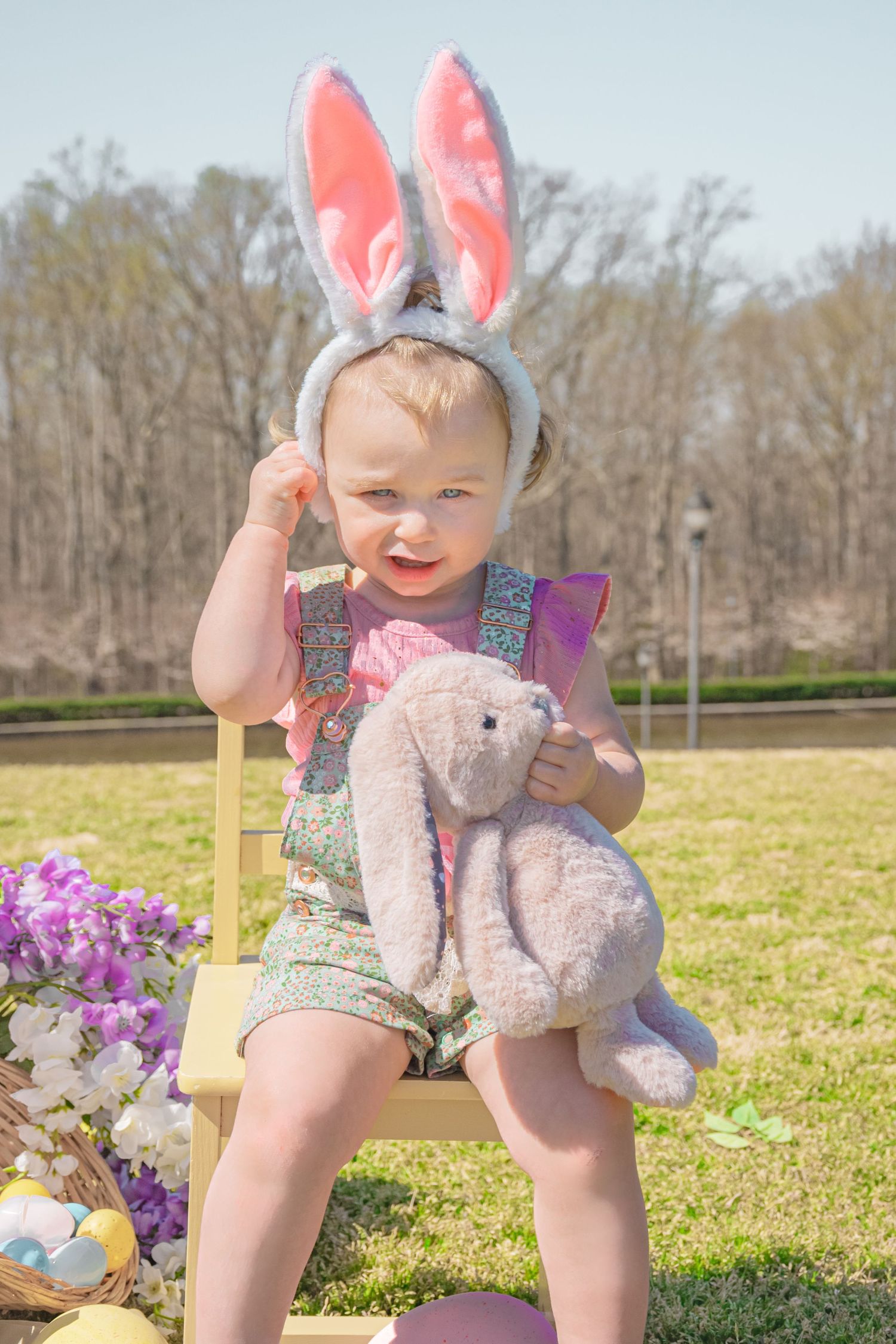 Cute toddler wearing bunny ears holding a stuffed rabbit toy while sitting outdoors during Easter celebration.