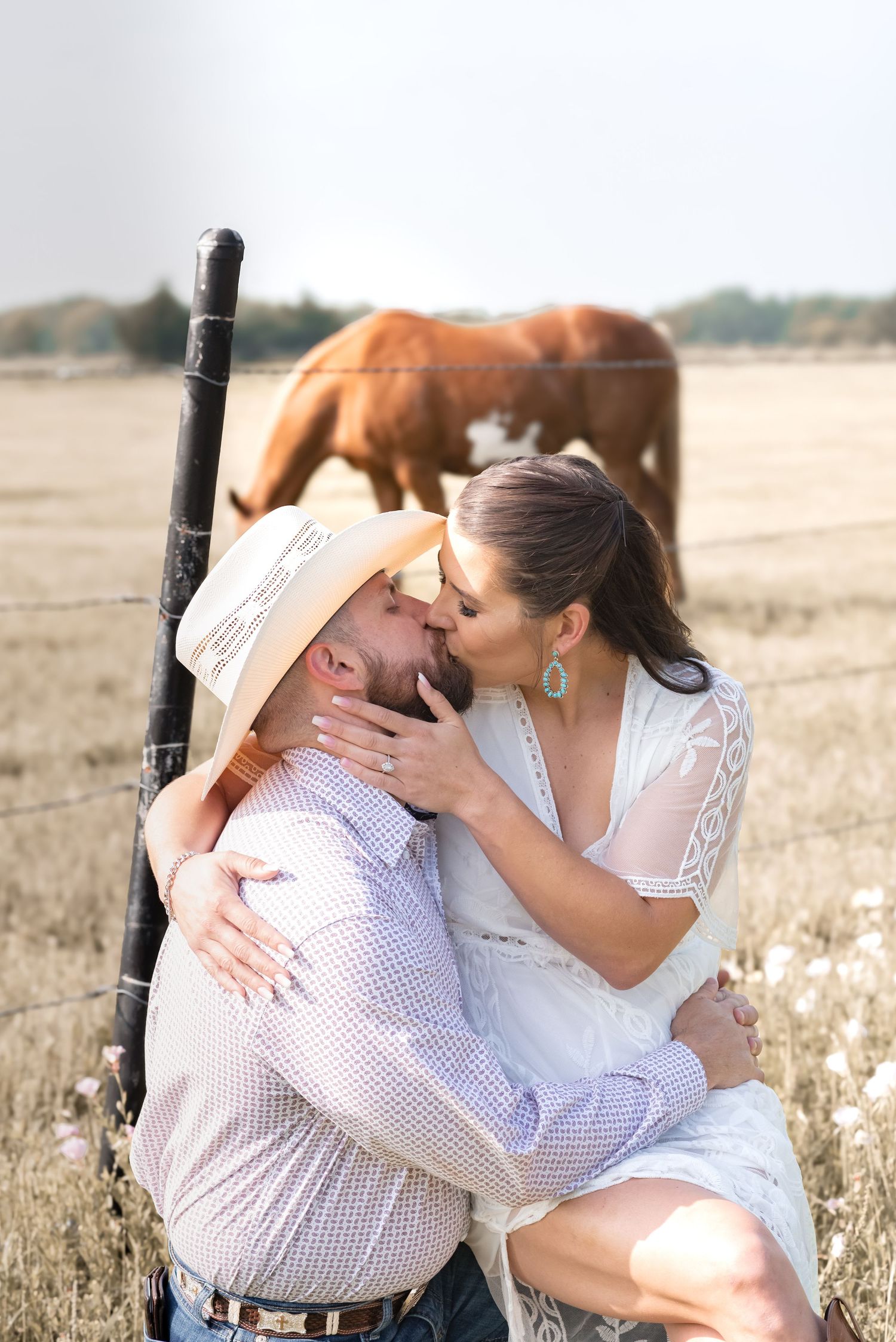 Couple sharing a romantic kiss near a fence post with a brown horse grazing in a golden field behind them.