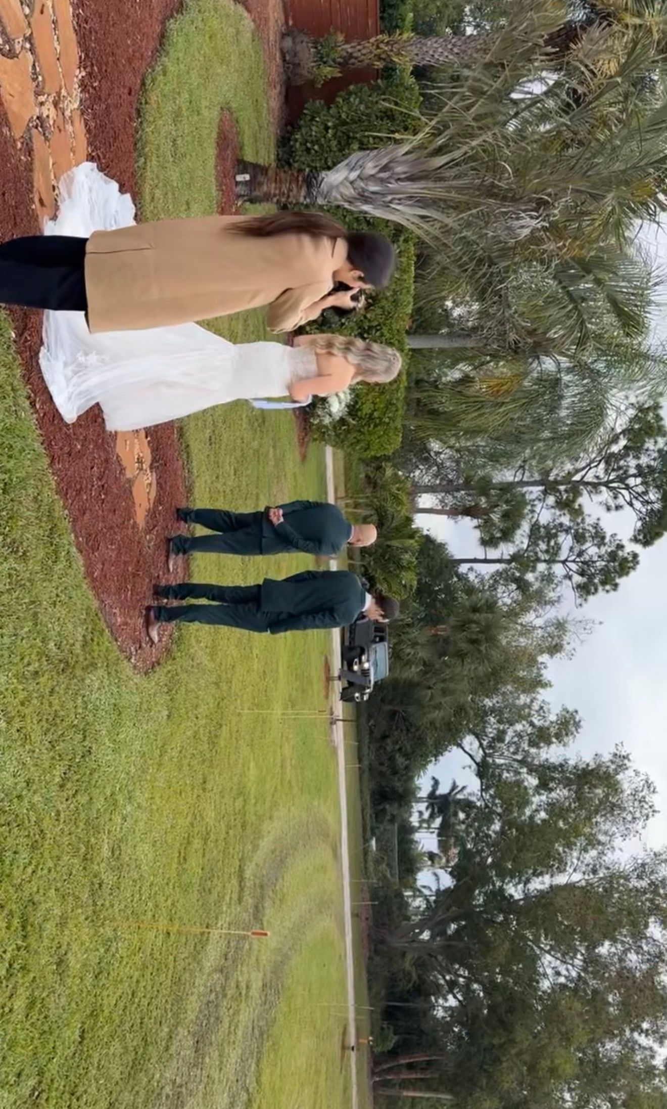 Behind-the-scenes wedding photography session with bride in white gown and groom on green lawn.