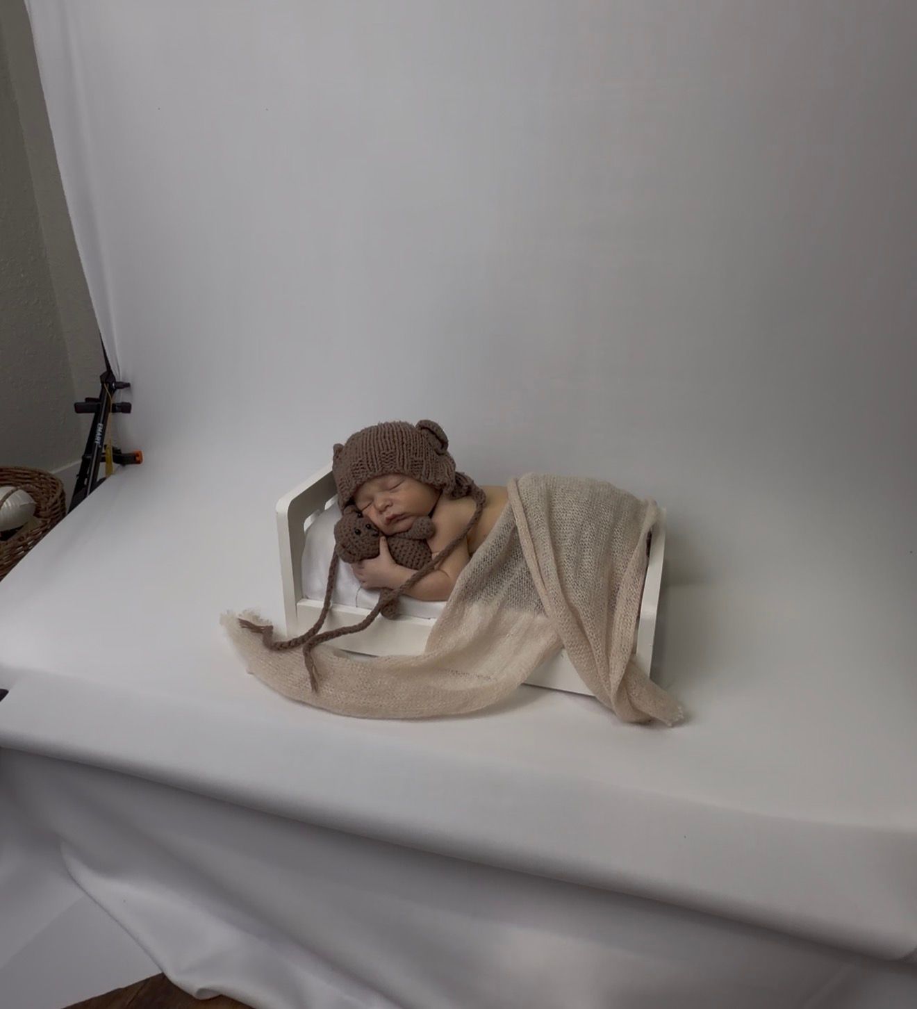 Sleeping newborn baby wrapped in beige blanket wearing brown bear bonnet during professional studio photoshoot.