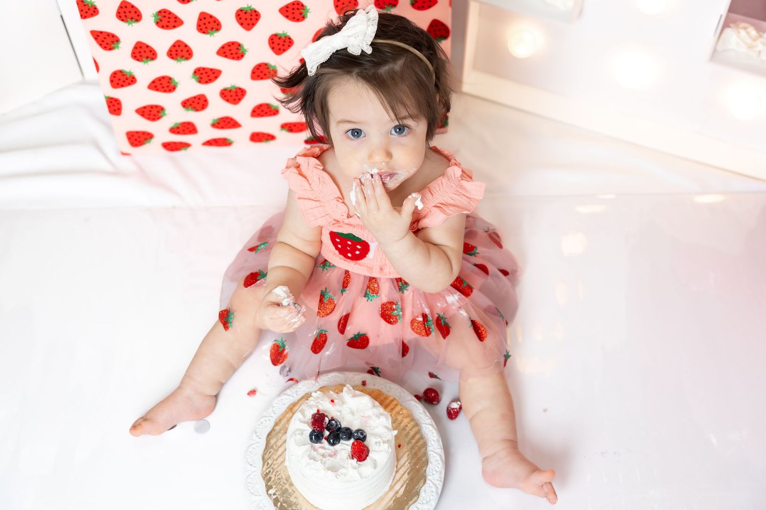A baby girl in a strawberry dress sits on a white floor, eating cake with berries at her first birthday smash.