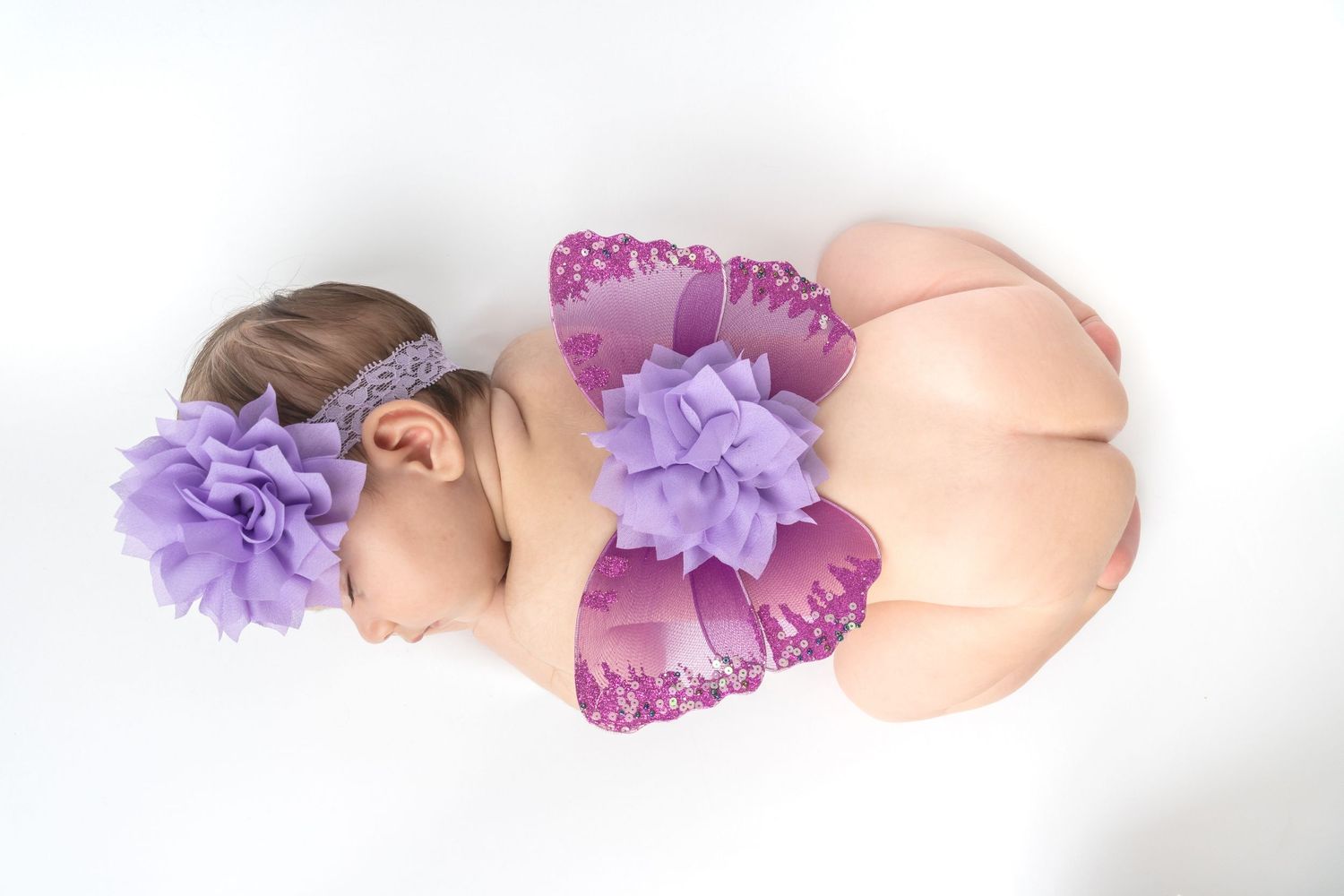 Newborn baby sleeping peacefully wearing purple butterfly wings and floral headband on white background.