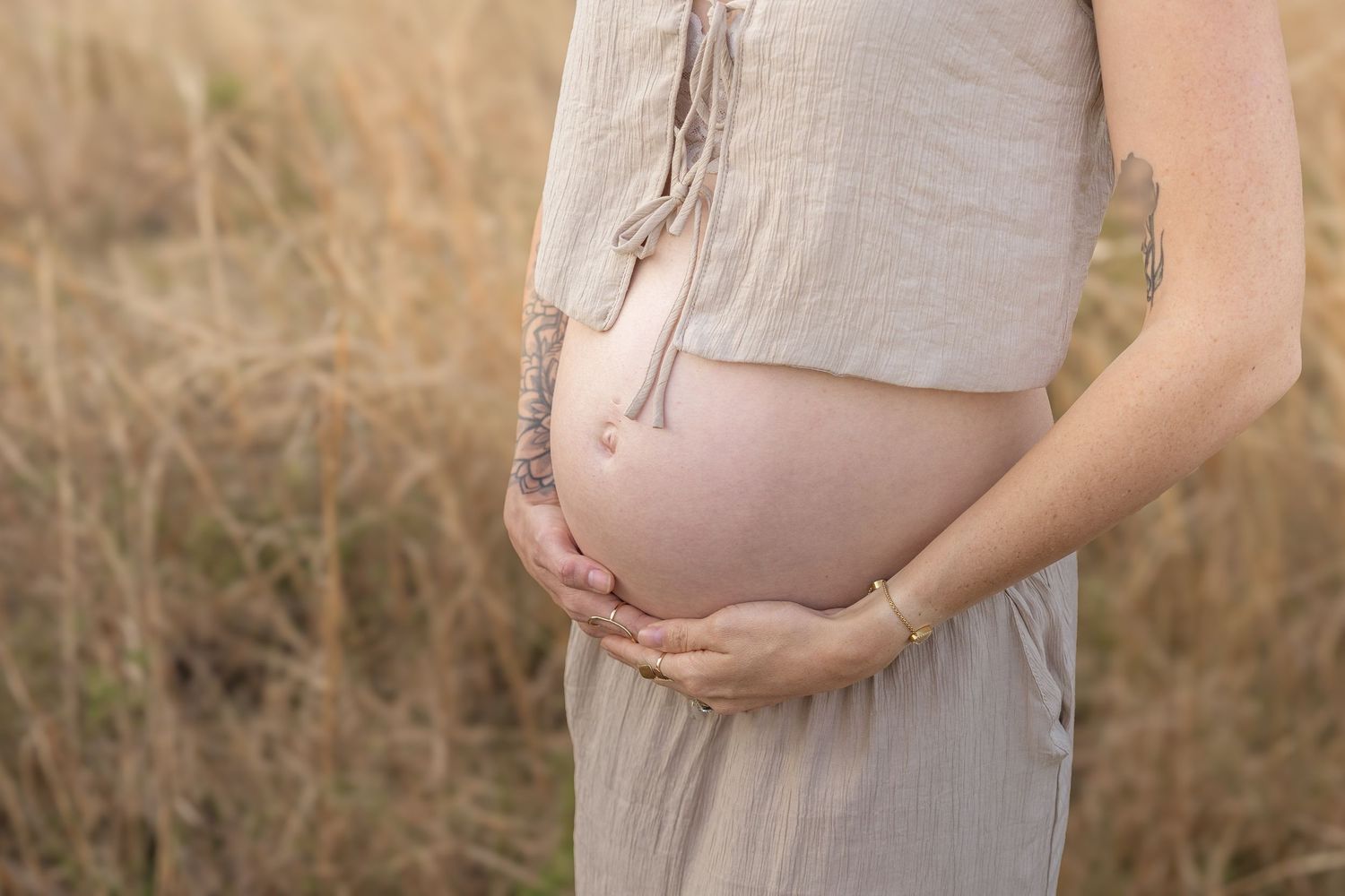 Pregnant woman in beige dress cradling bare baby bump outdoors in golden grass field, maternity photography.