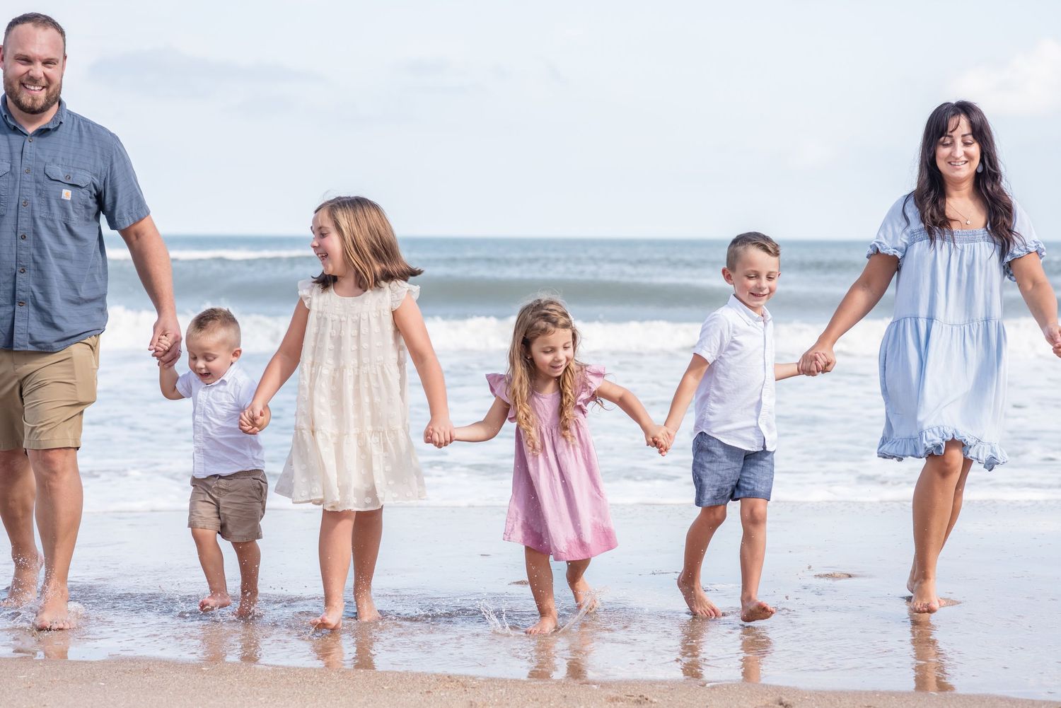 Family of six holding hands walking along a sandy beach shore with ocean waves in the background.