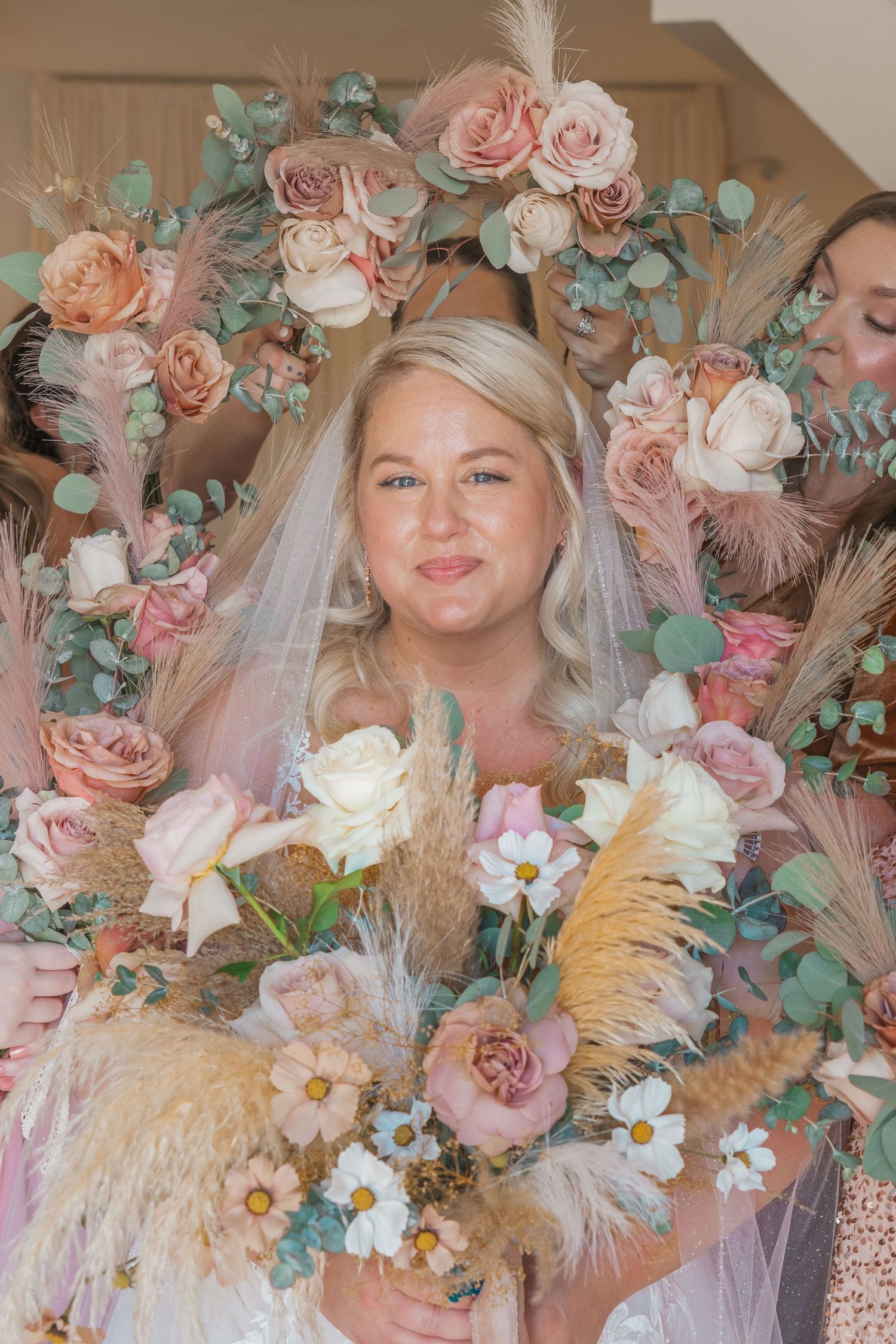 Bride holding a boho bouquet of blush roses and pampas grass surrounded by bridesmaids with floral hoops.