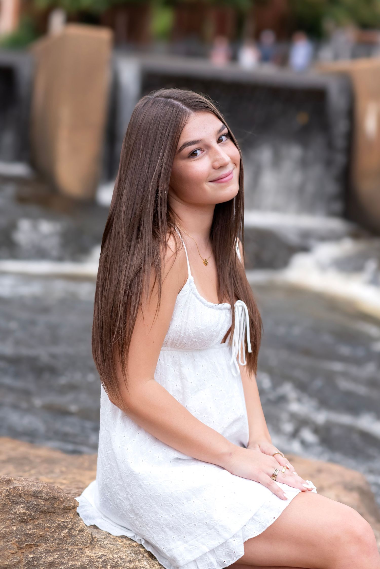 Young woman with long brown hair in white sundress sitting on rocks near a waterfall, smiling outdoors.
