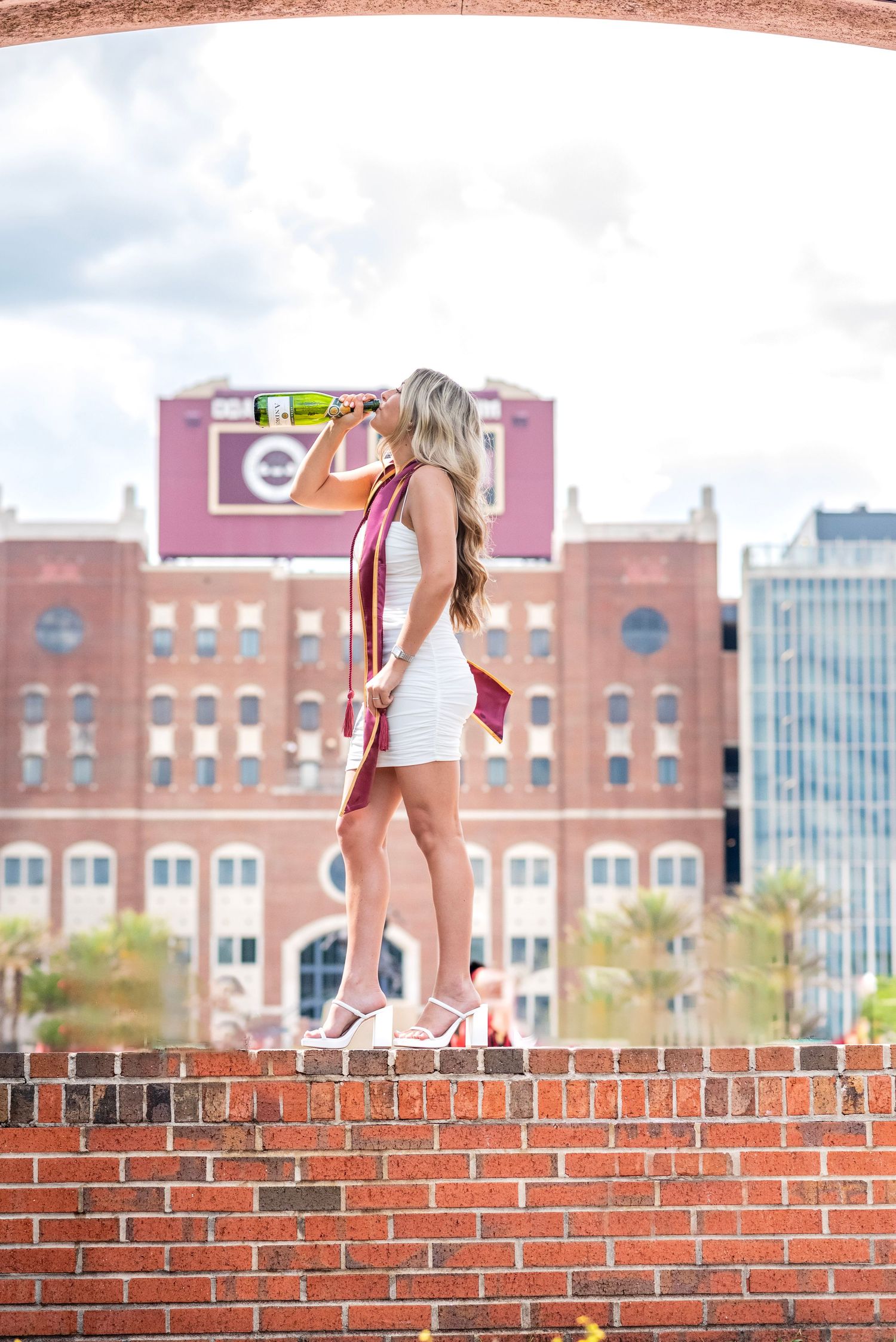 Young woman in white dress with maroon graduation sash stands on brick wall, drinking champagne with urban backdrop.