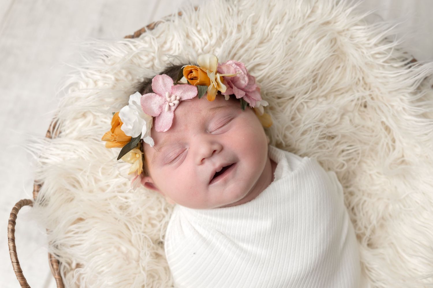 Sleeping newborn baby wrapped in white swaddle with floral crown resting in a basket with fluffy white fur.