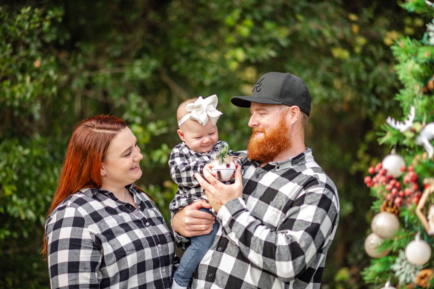 Family in matching plaid shirts holds baby near Christmas tree outdoors.